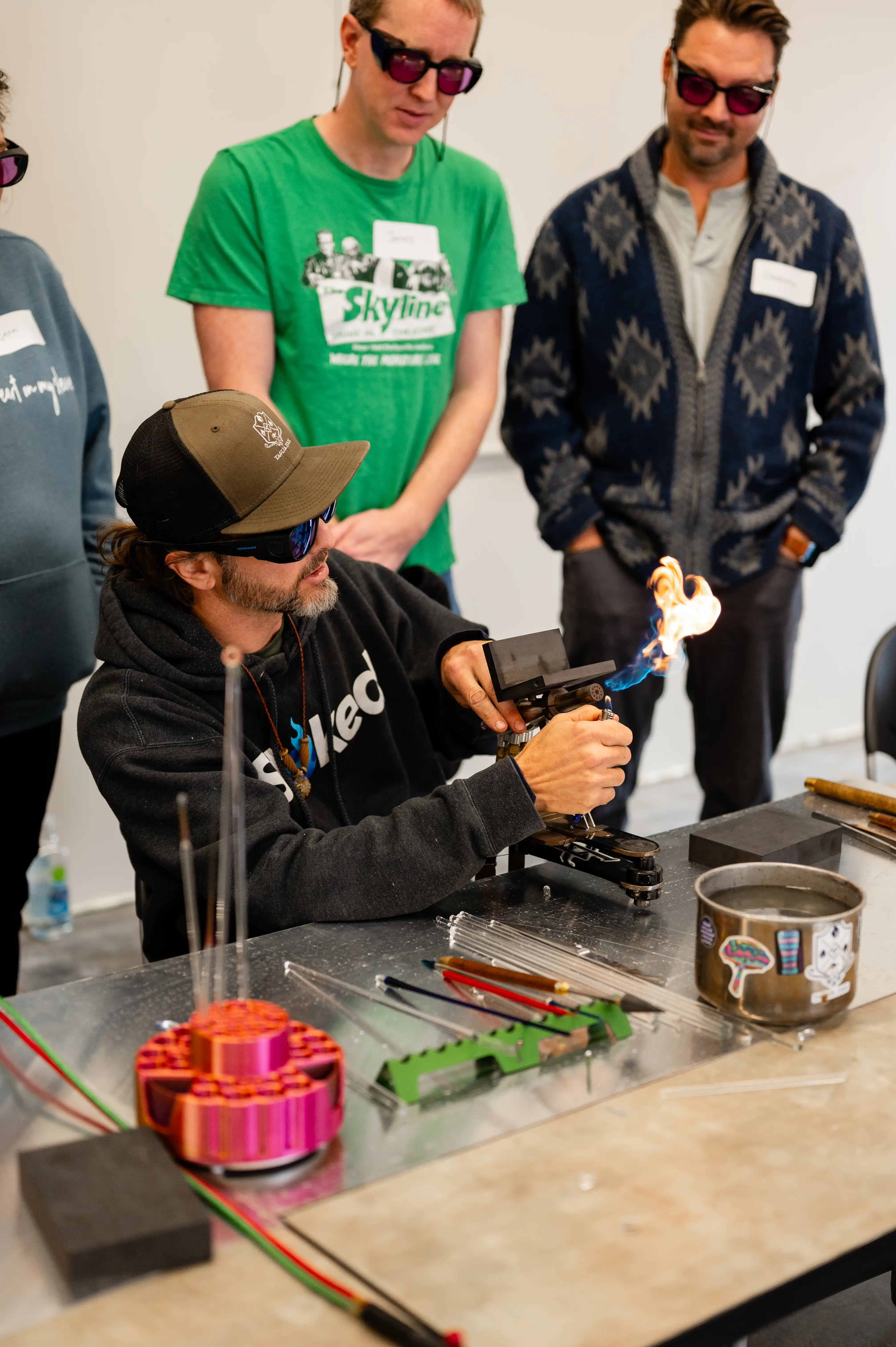 Person in a cap and sunglasses using a blowtorch to shape an object at a glassblowing workshop while onlookers watch.