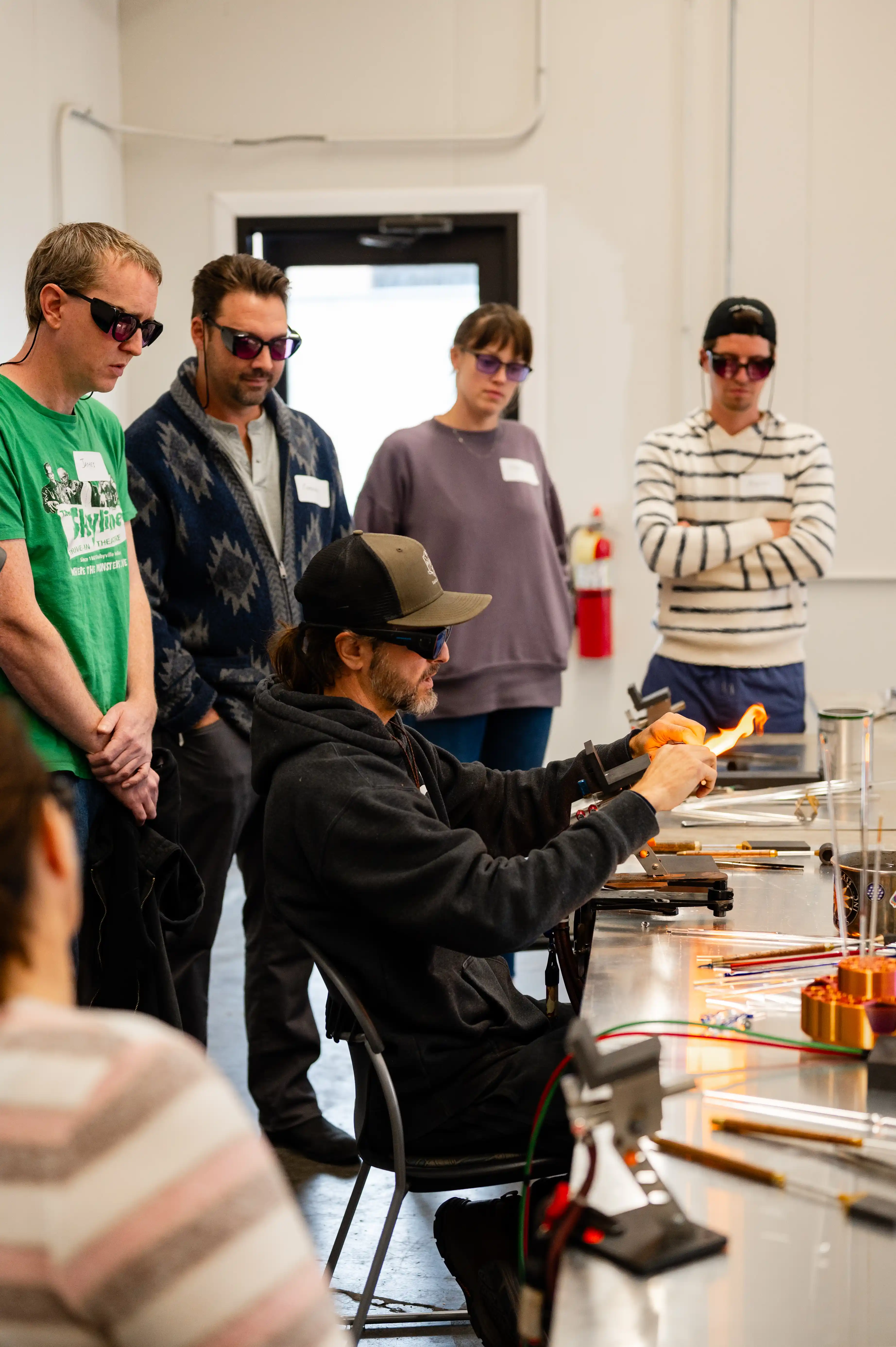 Glassblowing workshop with an instructor demonstrating techniques to a group of attentive students wearing protective eyewear.