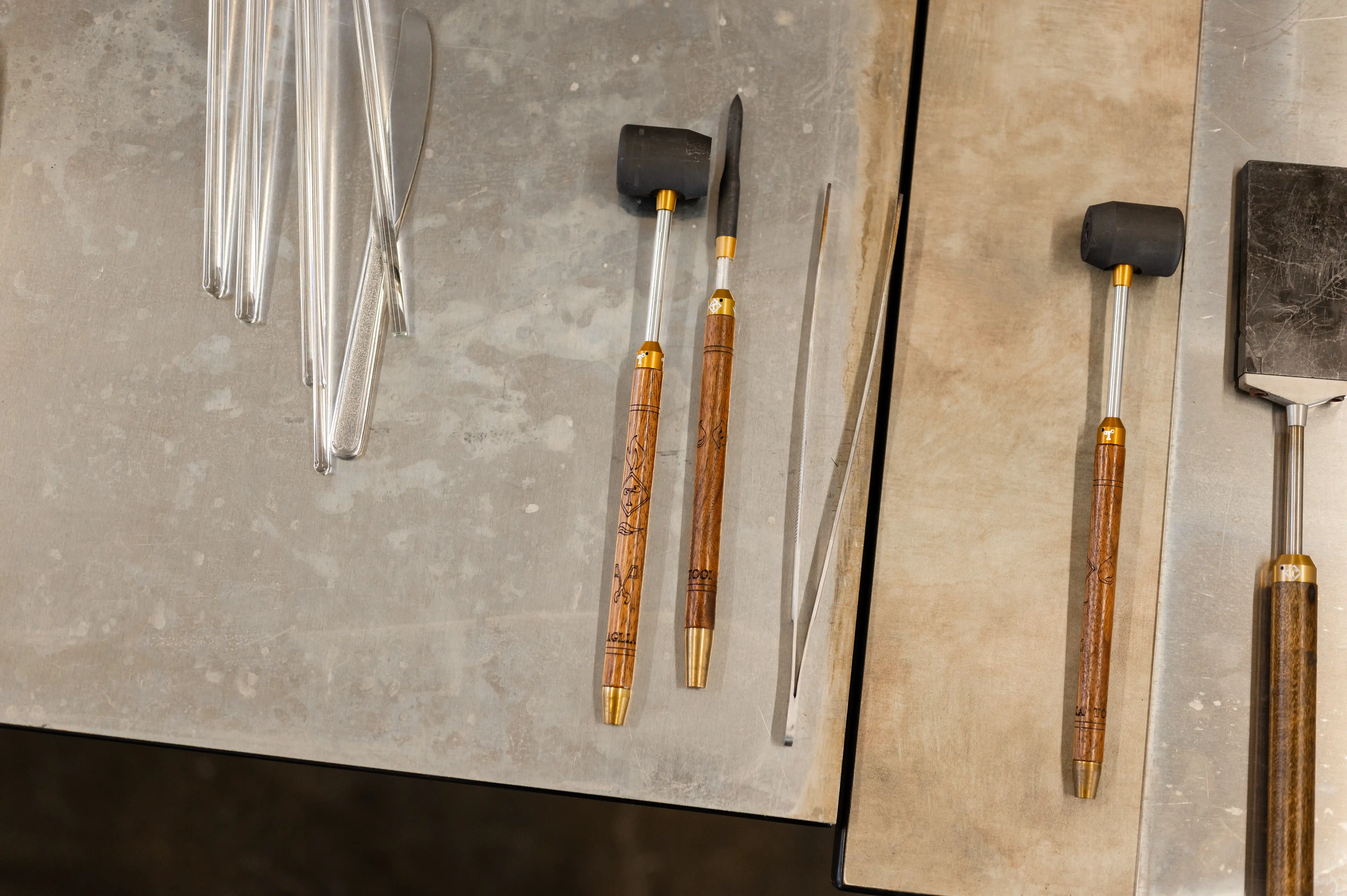 A collection of glassblowing tools including tweezers, jacks, and paddles organized on a steel workbench.