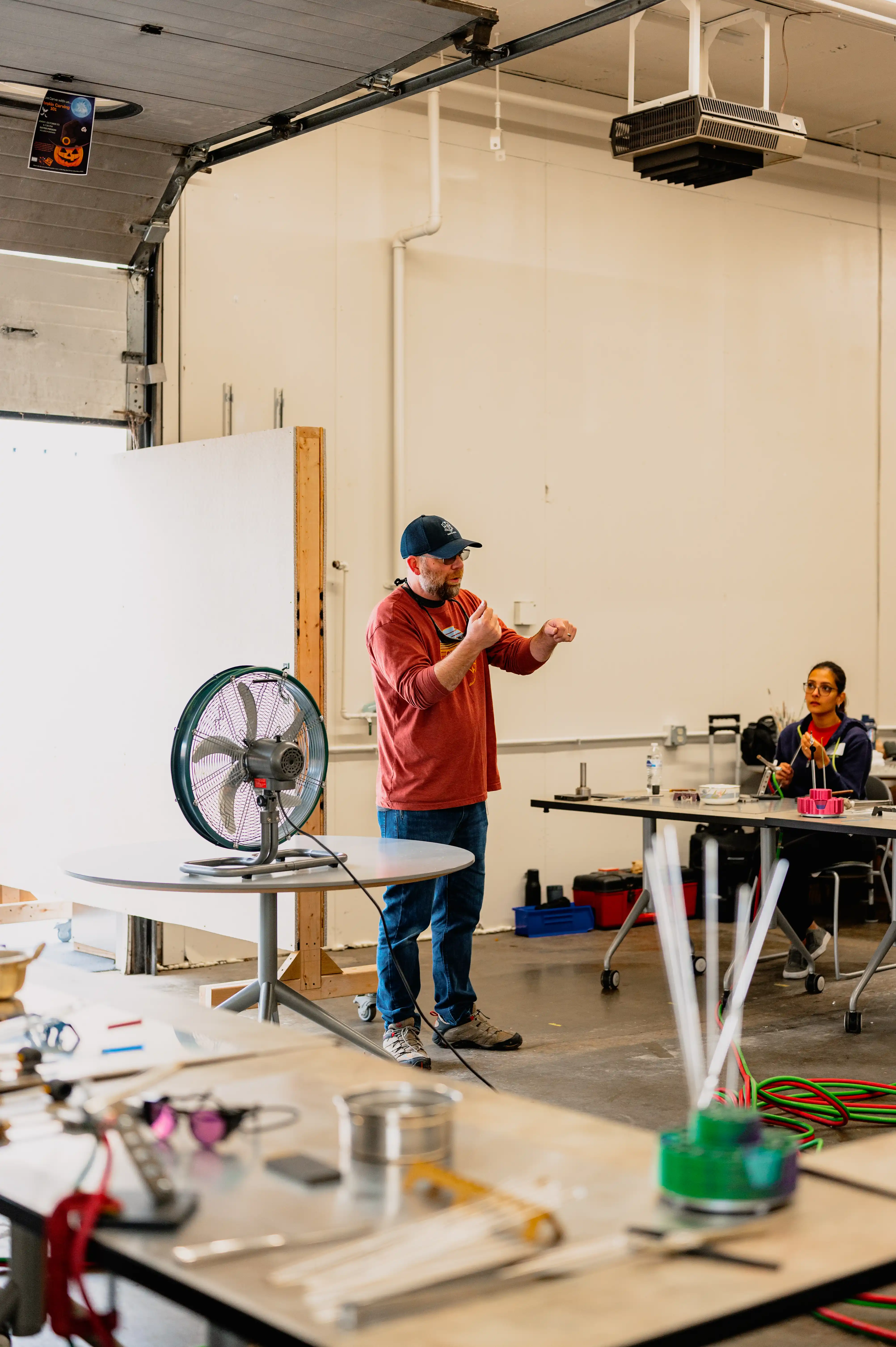 A man in a baseball cap and red sweatshirt stands speaking with a coffee cup in hand in an industrial workshop, while a woman seated at a workbench focuses on an electronic device.