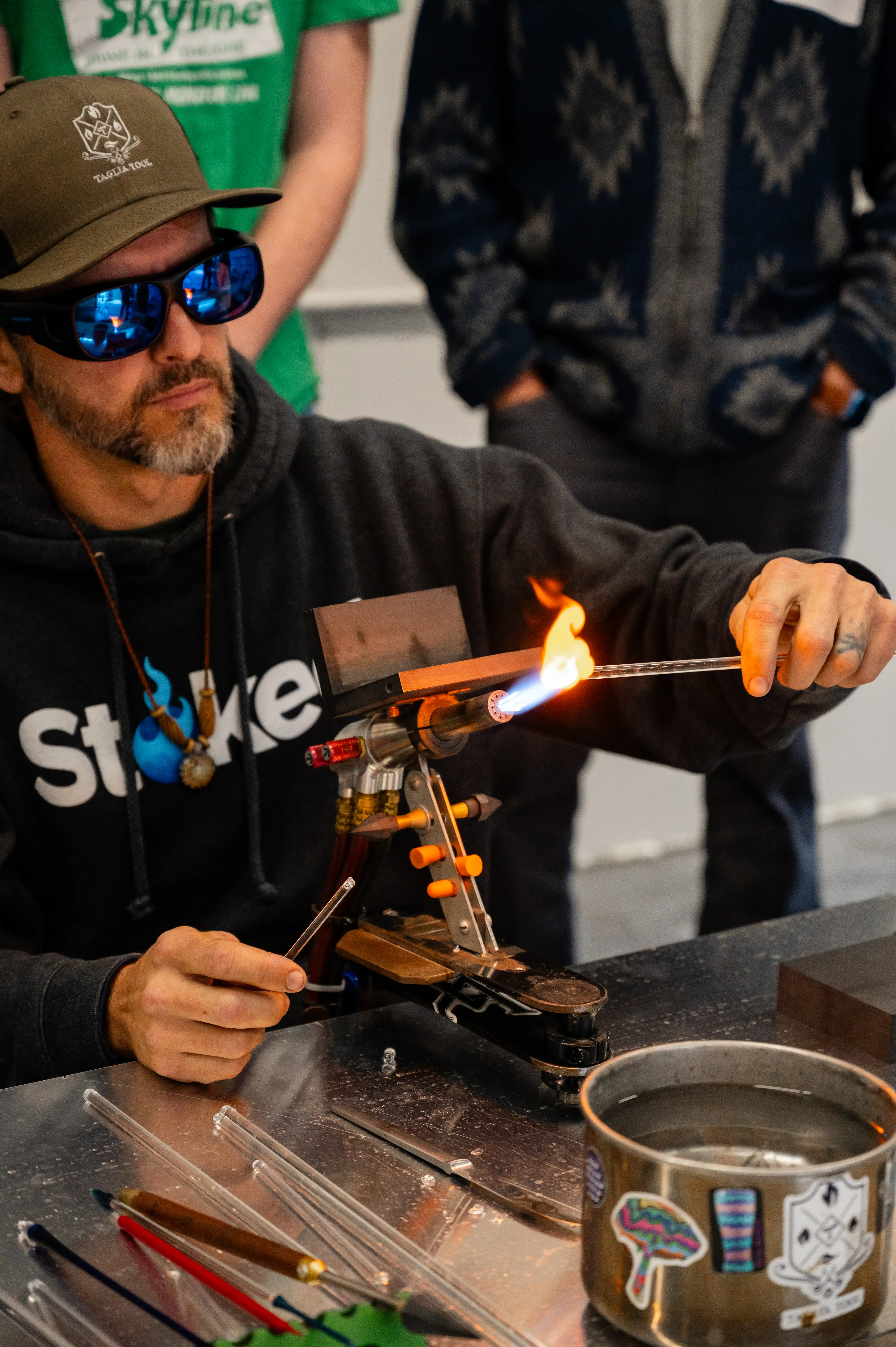 Glassblower using a torch to shape a piece of glass, wearing sunglasses and a hooded sweatshirt, with tools on the table in front.