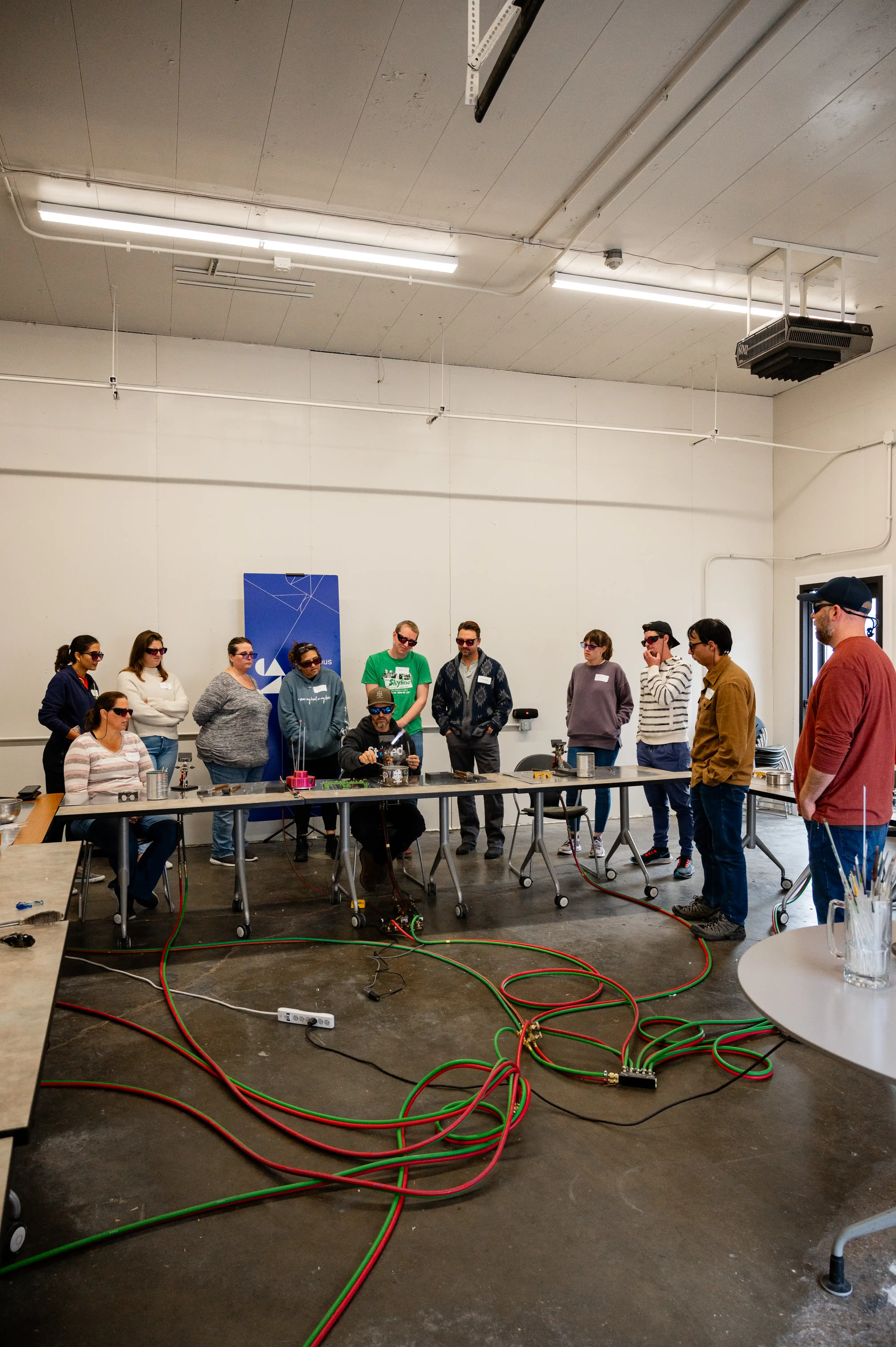 Group of people standing around a table in a workshop environment observing a demonstration or lecture.