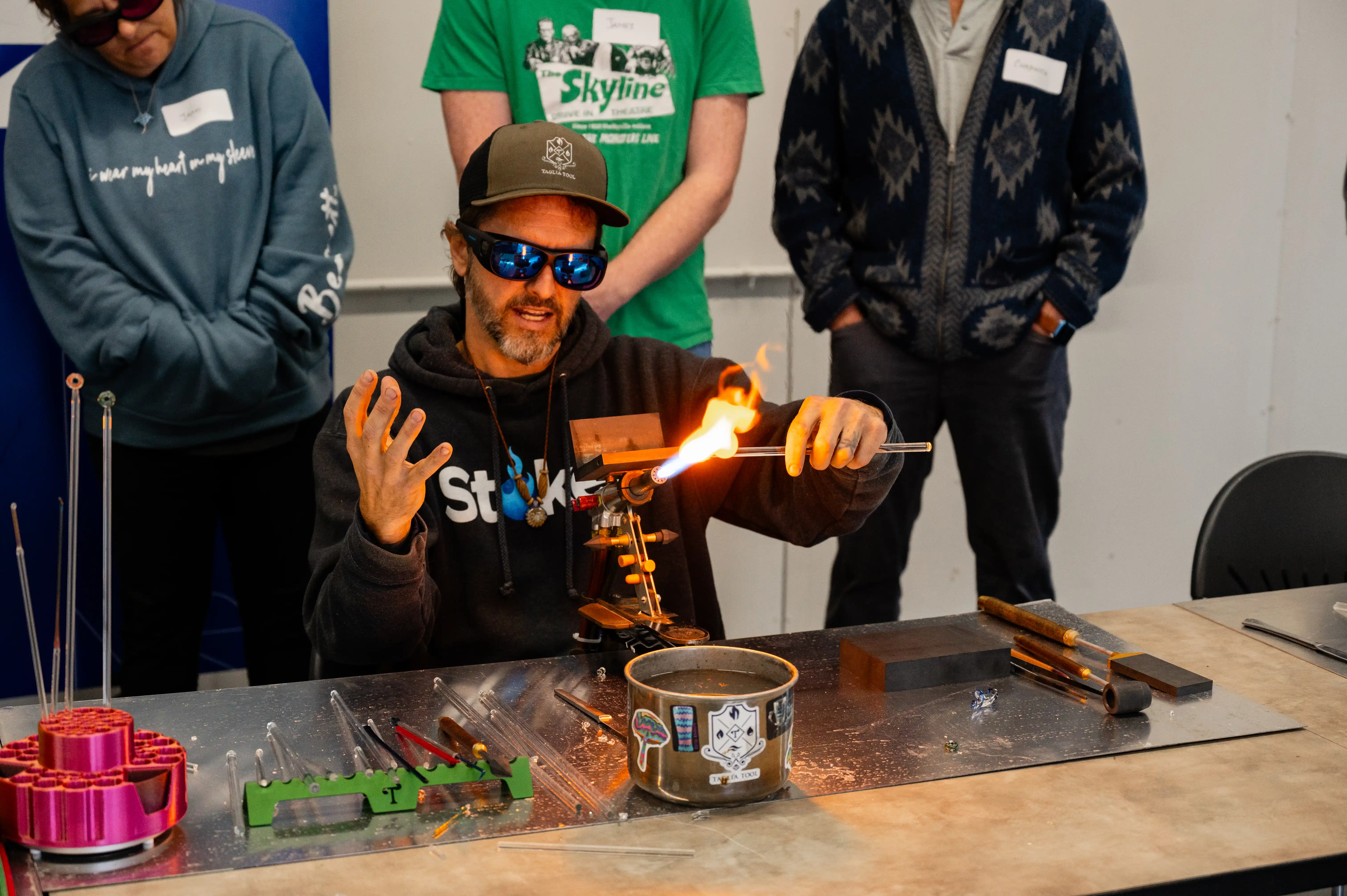 Glassblower demonstrating techniques with a torch and molten glass in a workshop setting, surrounded by various glassblowing tools, with onlookers wearing glasses.