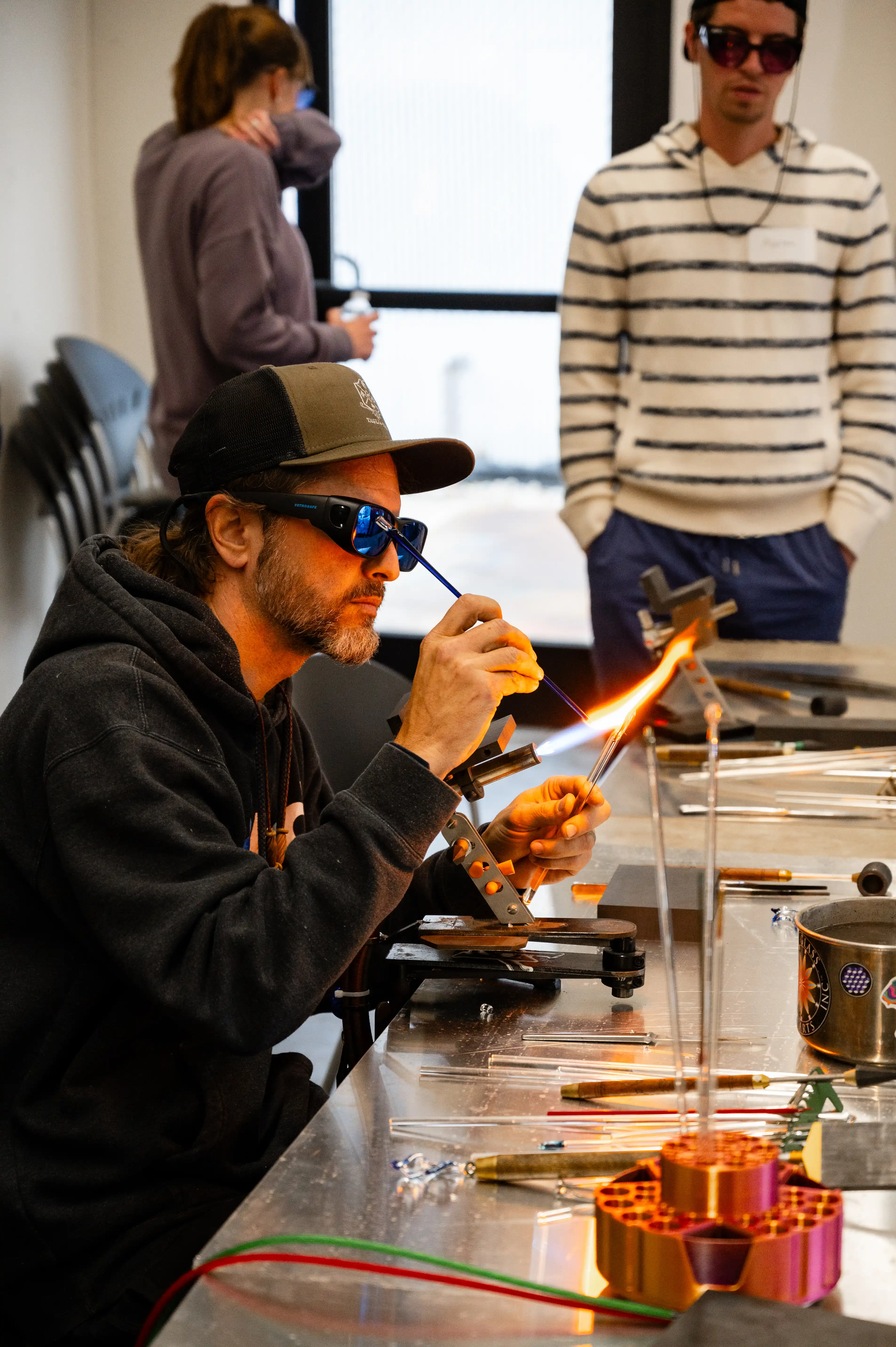 Glassblower creating glass art using a torch in a workshop with observers in the background.