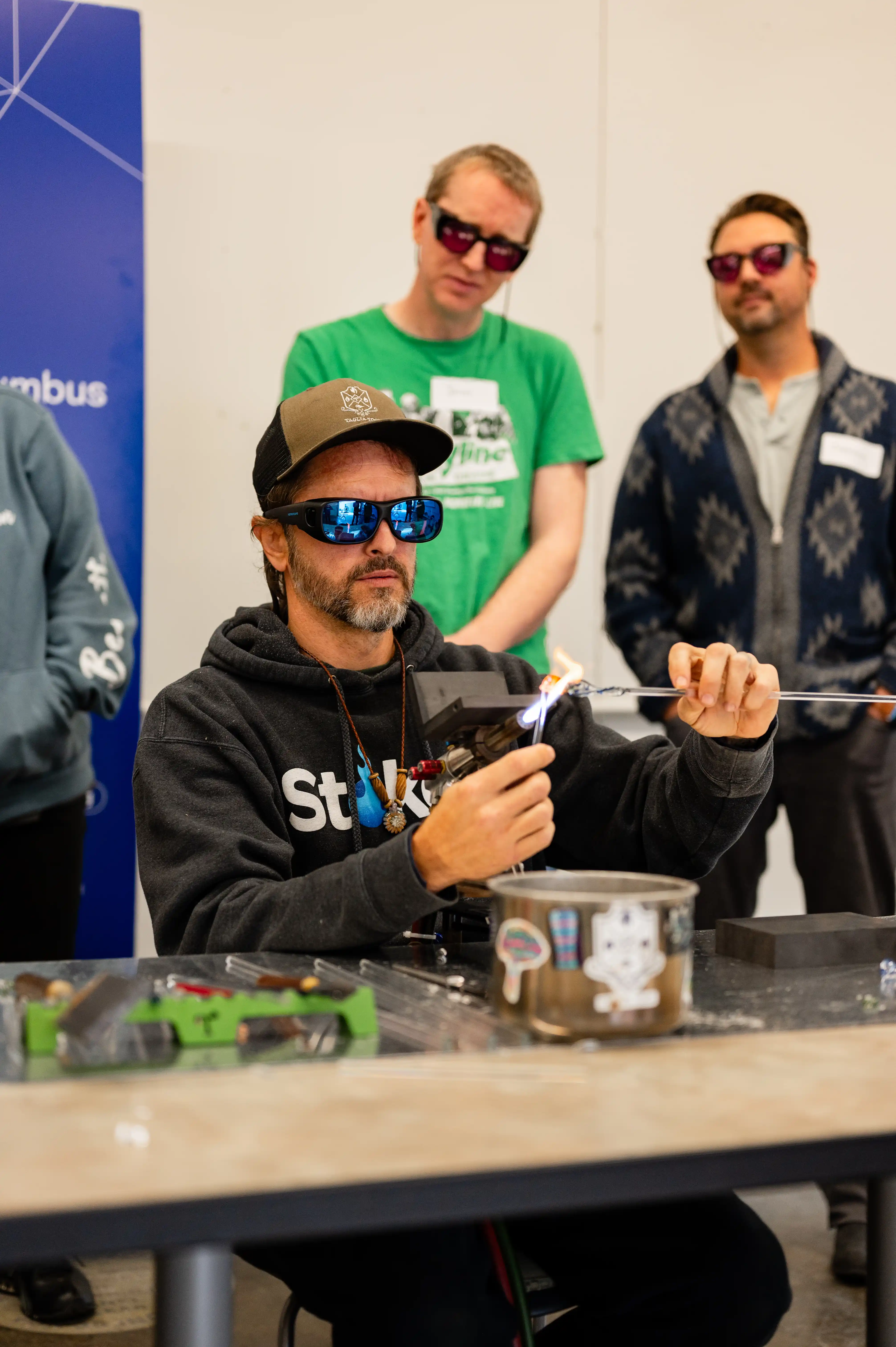 Man wearing sunglasses and a hat doing glassblowing with a torch, with two observers wearing protective eyewear in the background.