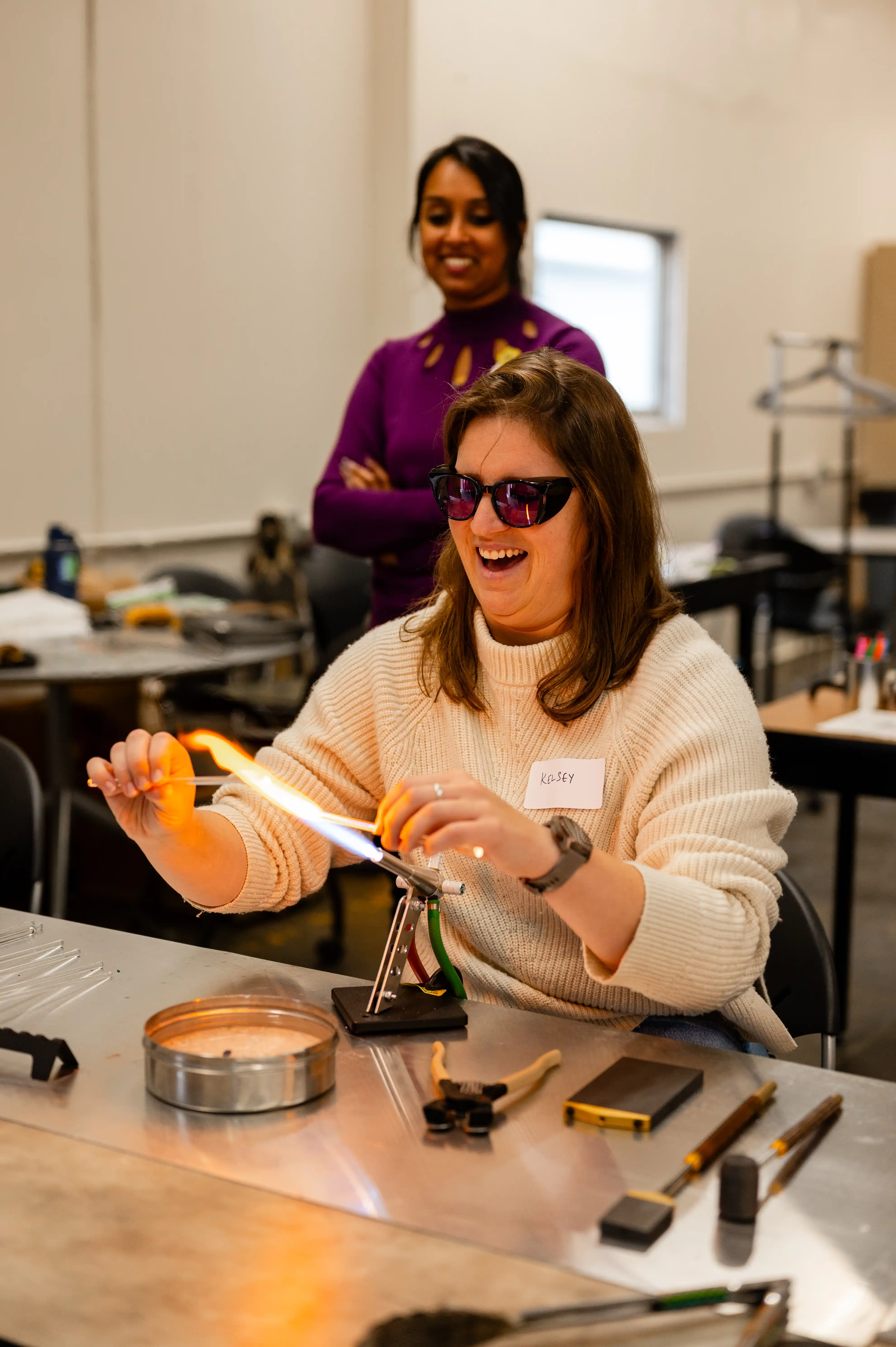 Woman in a cream sweater and sunglasses laughing while shaping a glass piece with a torch as another woman looks on in a workshop environment.