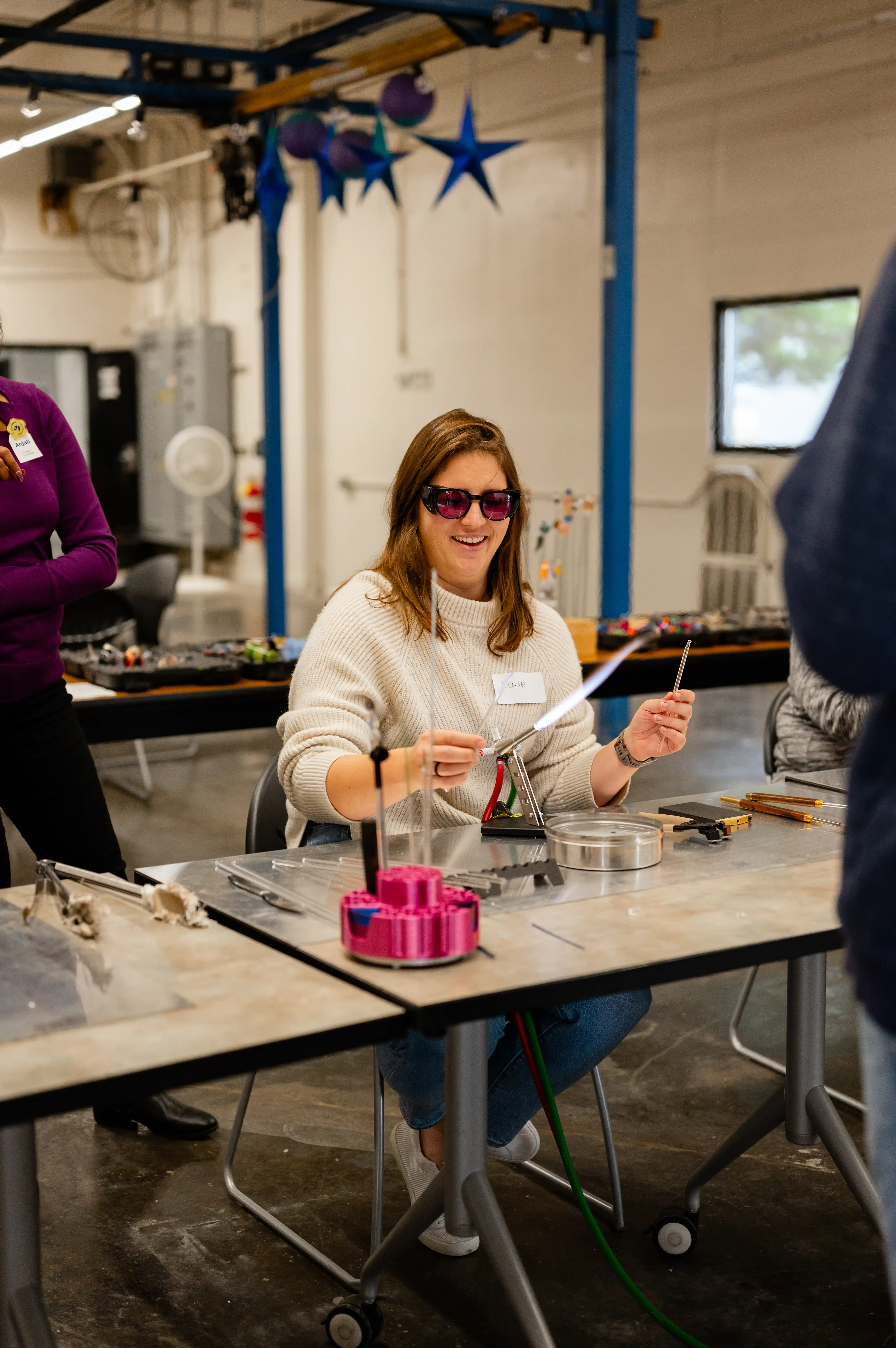 Woman wearing safety glasses sitting at a workshop table, smiling and holding a tool, with other participants and equipment around her.