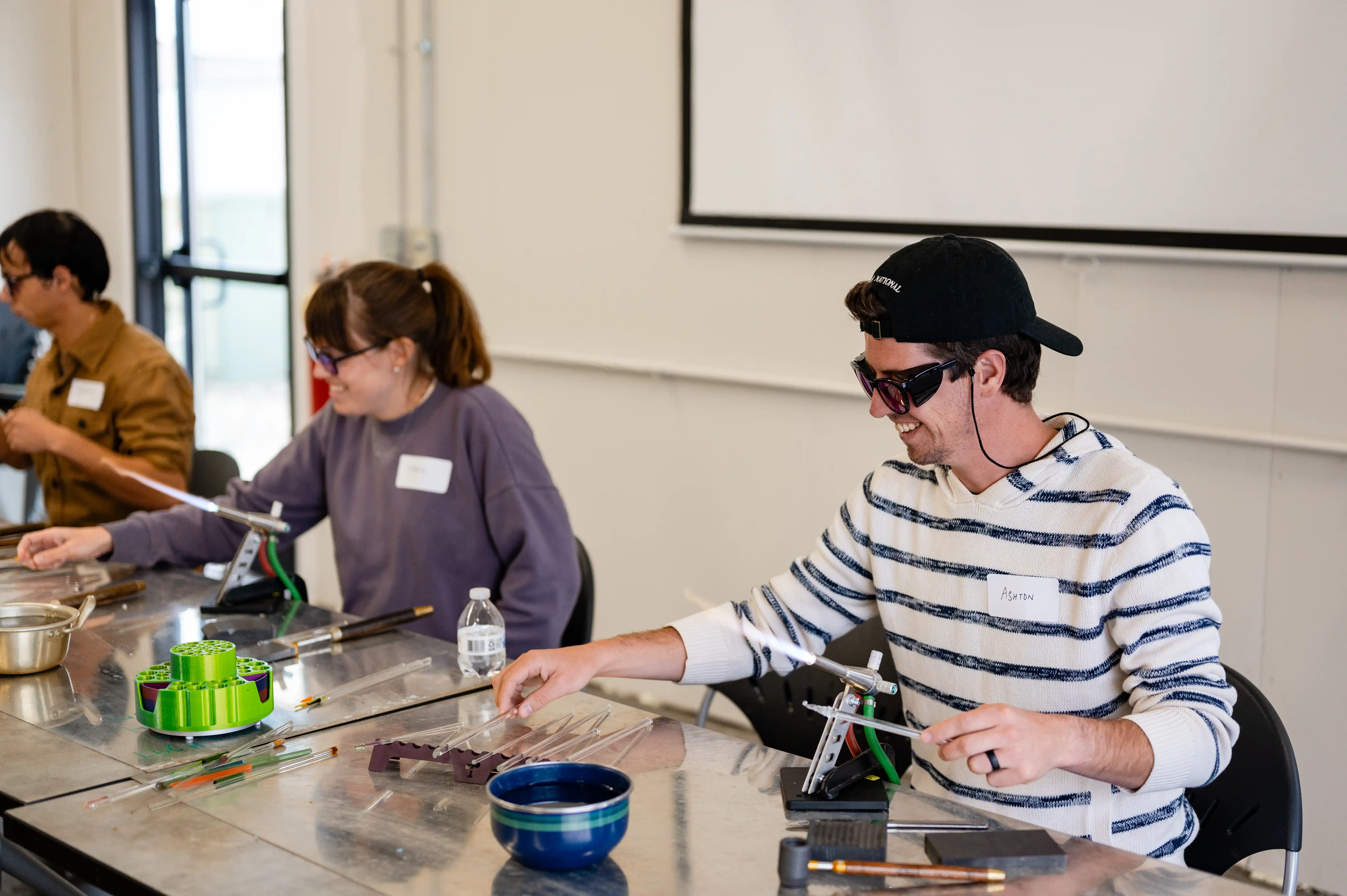 People engaging in a glassworking workshop, smiling as they manipulate various tools and colored glass rods at a well-lit table.