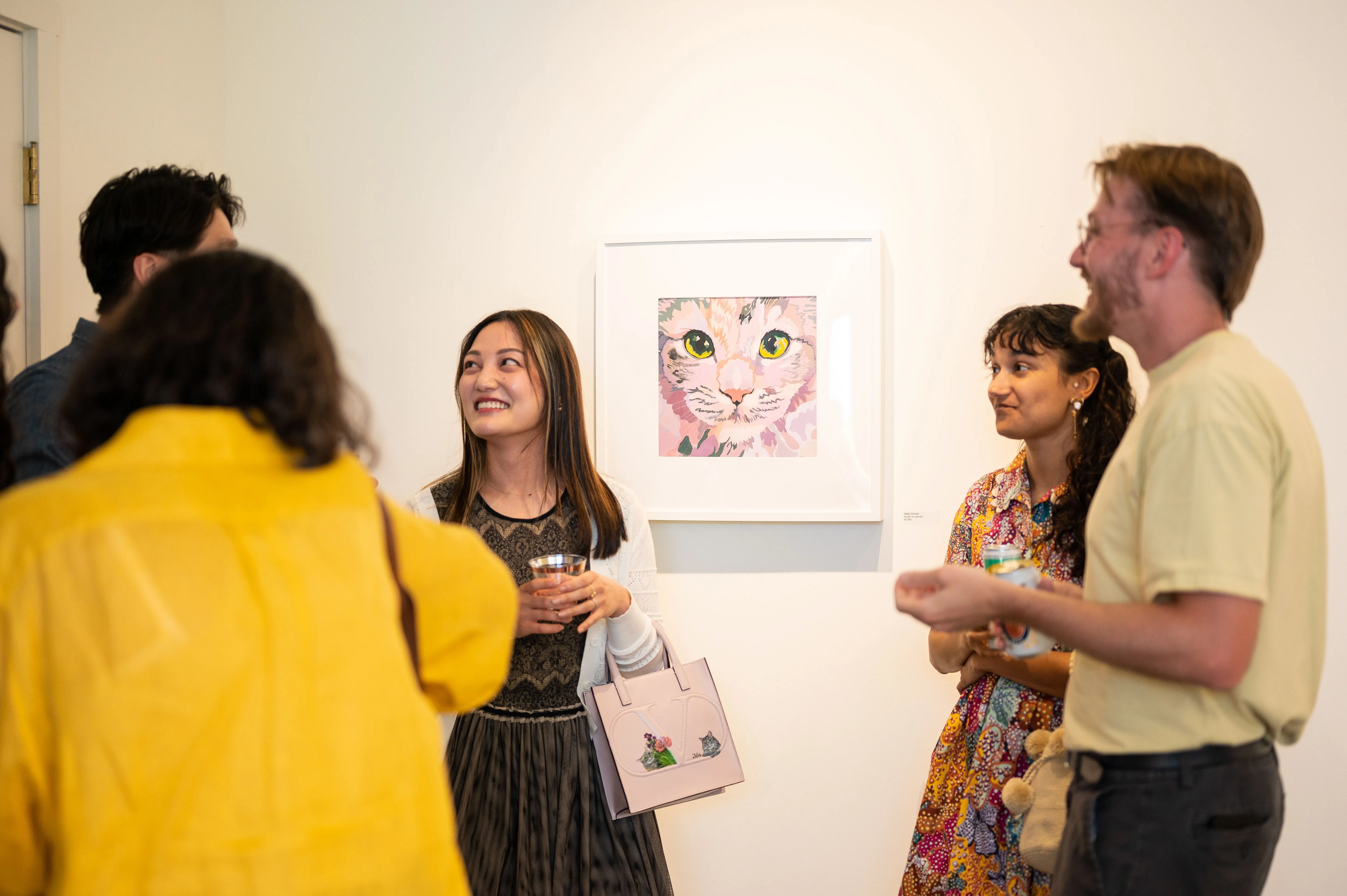 Group of people engaging in conversation at an art gallery, with a colorful artwork of a cat on the wall in the background.