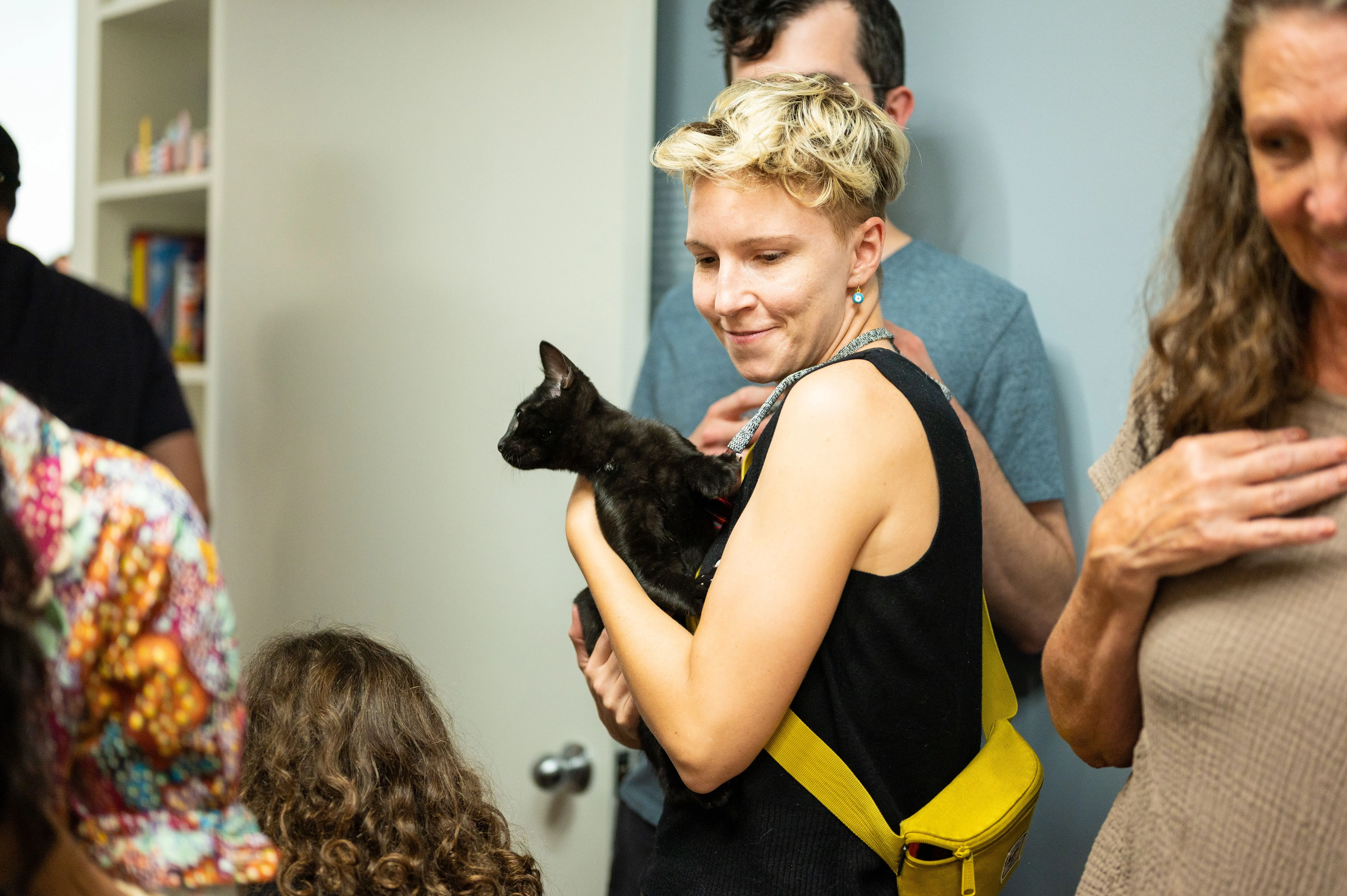 Woman smiling and holding a black cat in a room with other people.