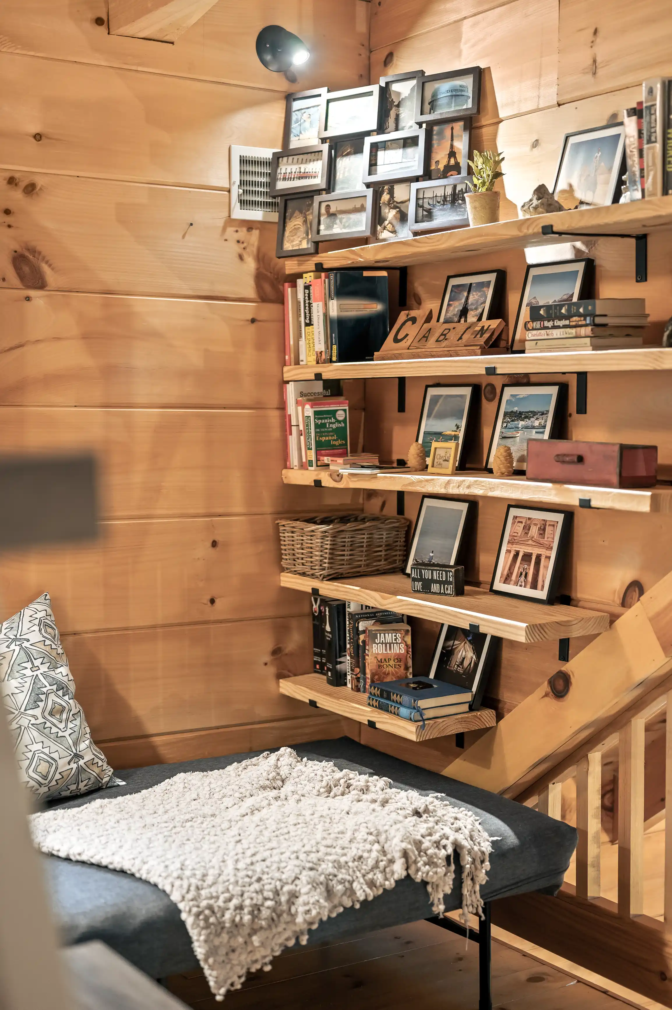 A cozy corner with a bench and a white cushion, adjacent to wooden shelves filled with books, framed pictures, and decorative items, with warm lighting in a cabin interior.