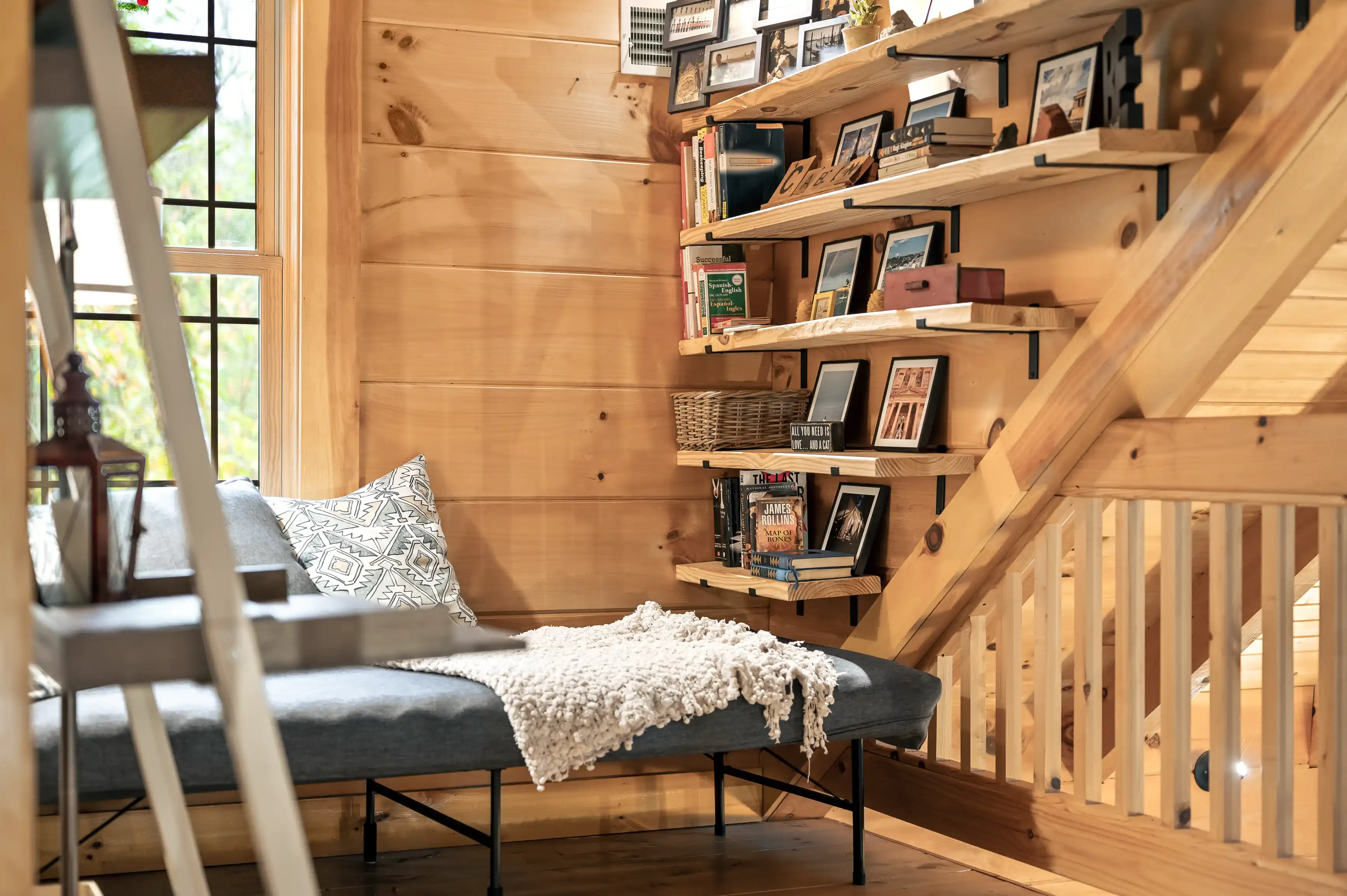 Cozy wooden cabin interior with a staircase lined with bookshelves full of books and framed photos, and a window seat with cushions and a throw blanket.