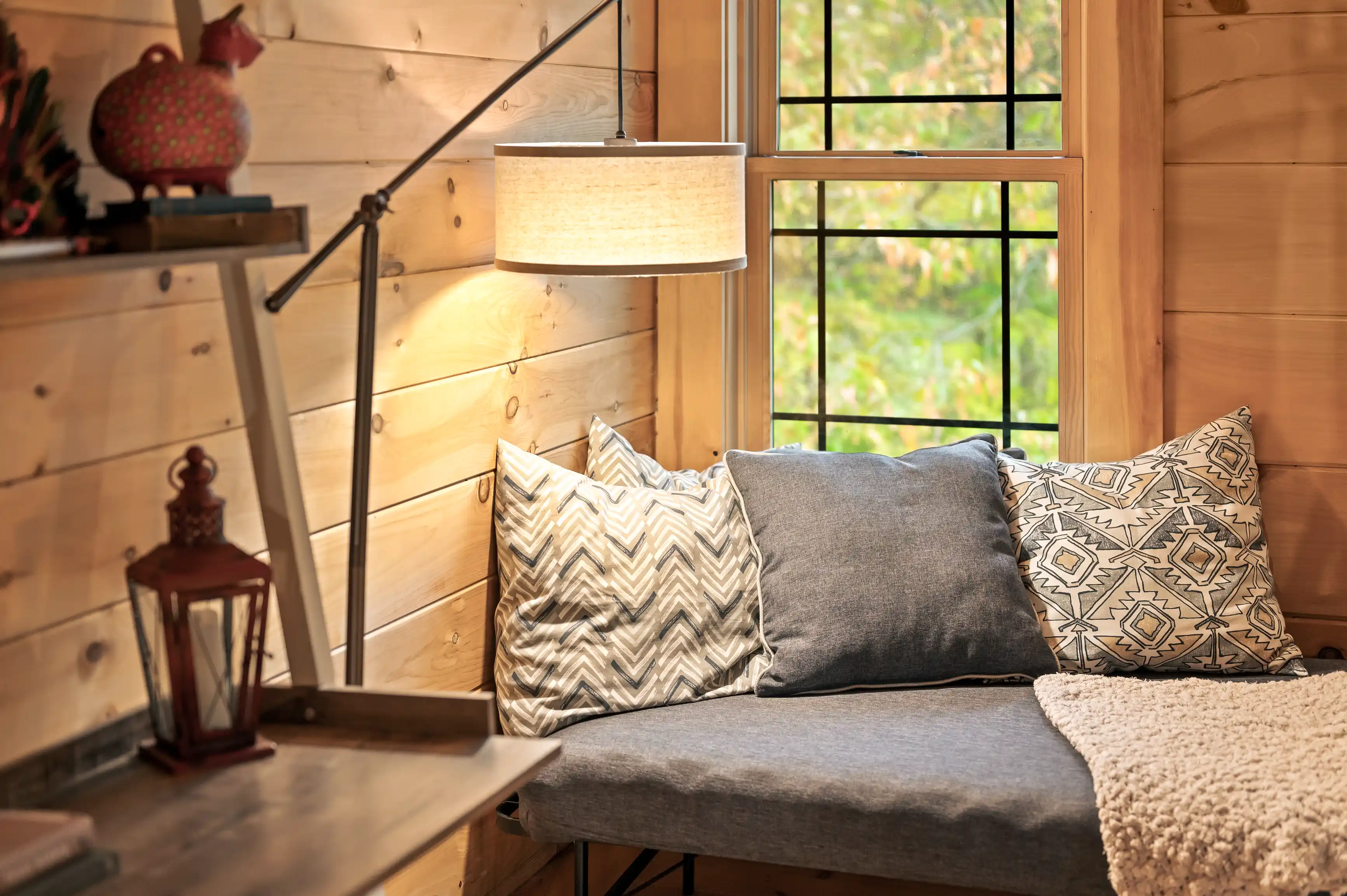 Cozy wooden cabin interior with a bench adorned with cushions, a lit floor lamp, a rustic lantern, and a window overlooking greenery.