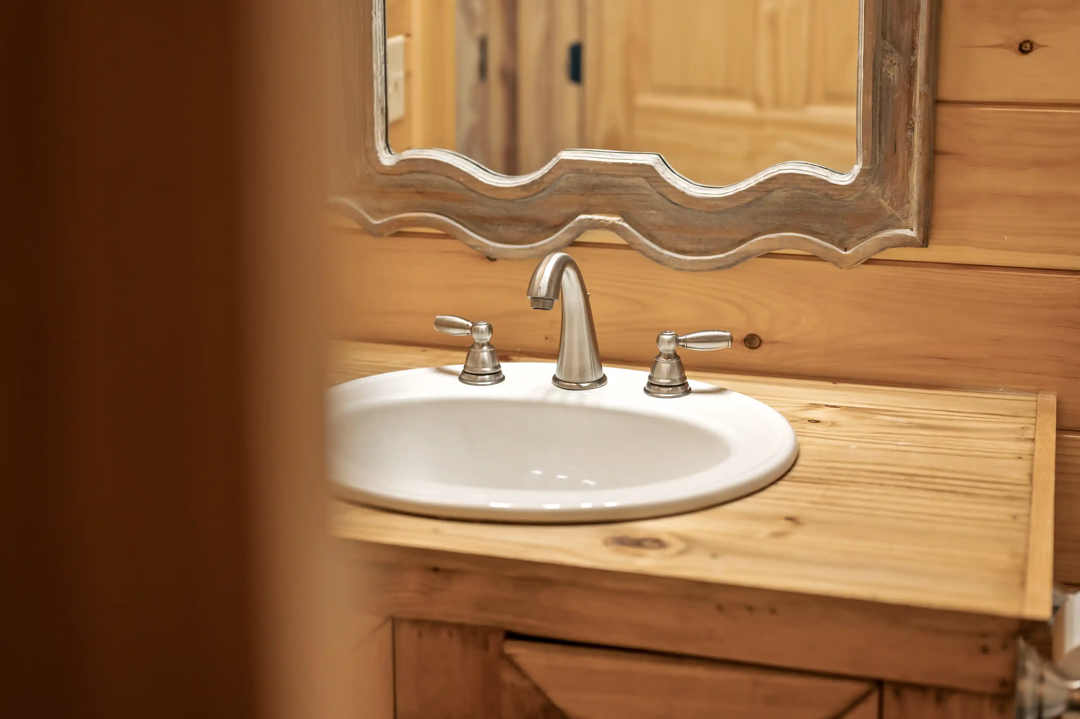 Rustic wooden bathroom interior with a white ceramic sink, silver faucet, and a decorative mirror on a wooden wall.