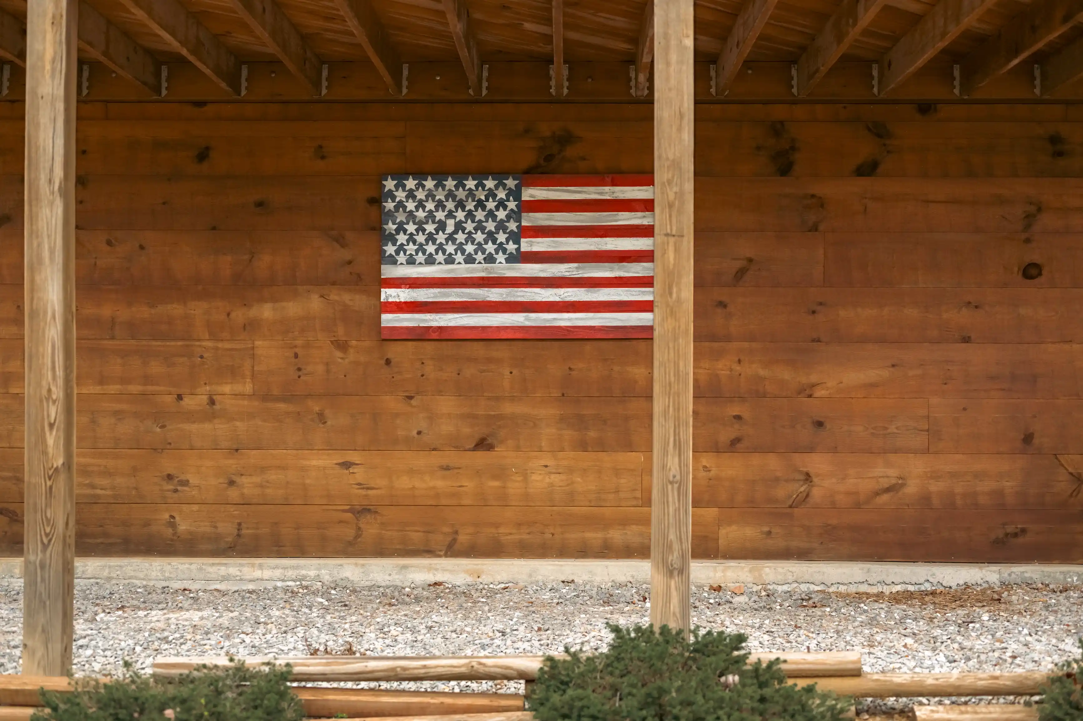 An American flag hanging on a wooden wall inside a covered area with wooden beams and a gravel ground.