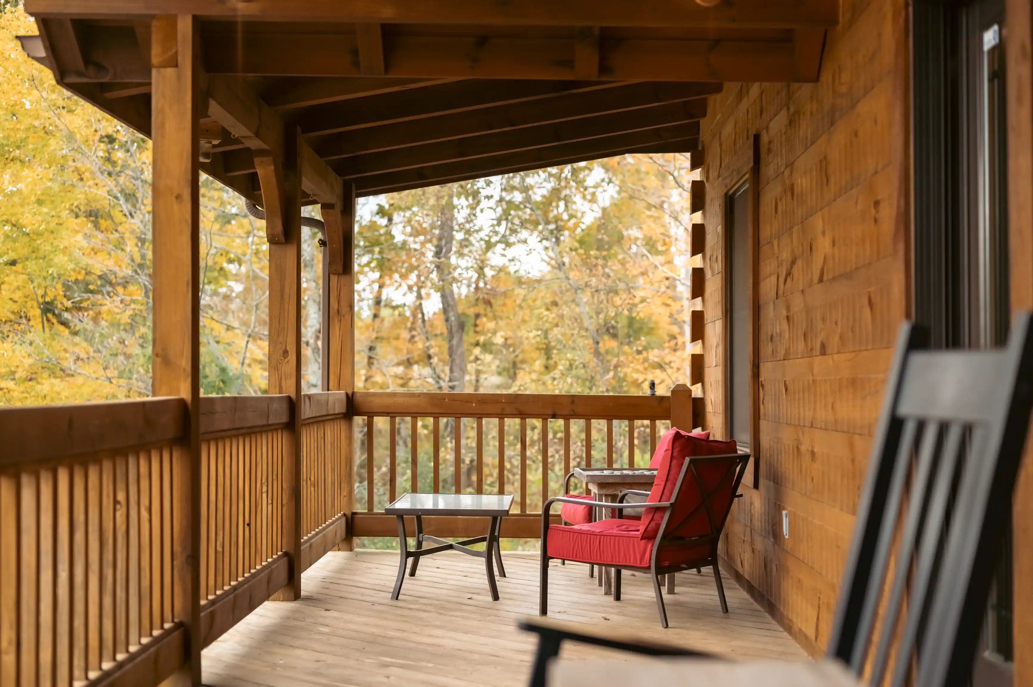 Cozy wooden cabin porch with a red chair and a table overlooking autumn trees.