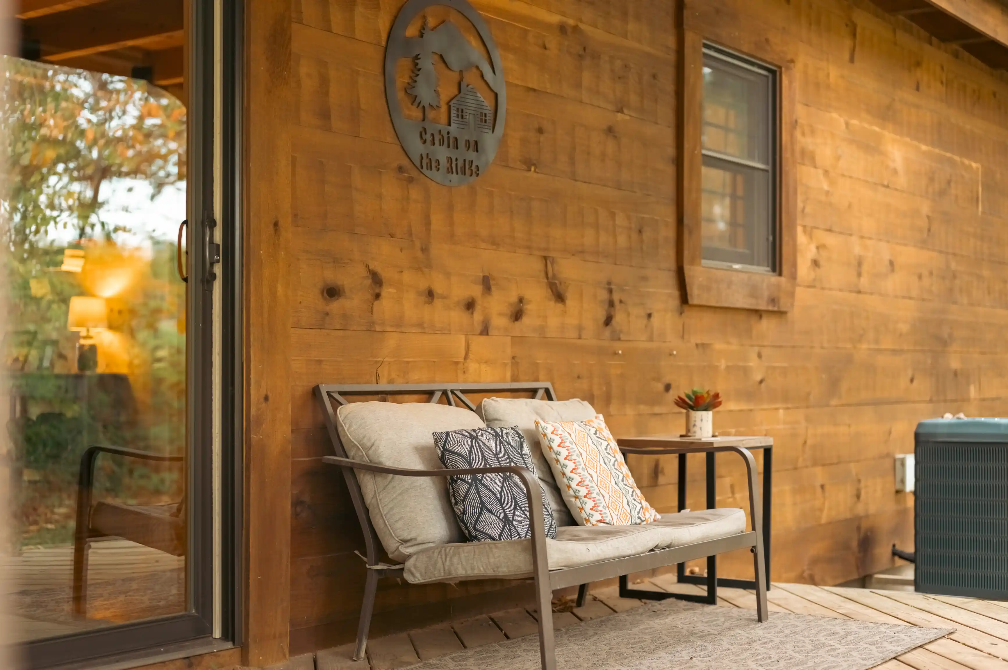 A cozy wooden cabin's porch with a metal bench, decorative pillows, and a sign that reads "Cabin on the Ridge".