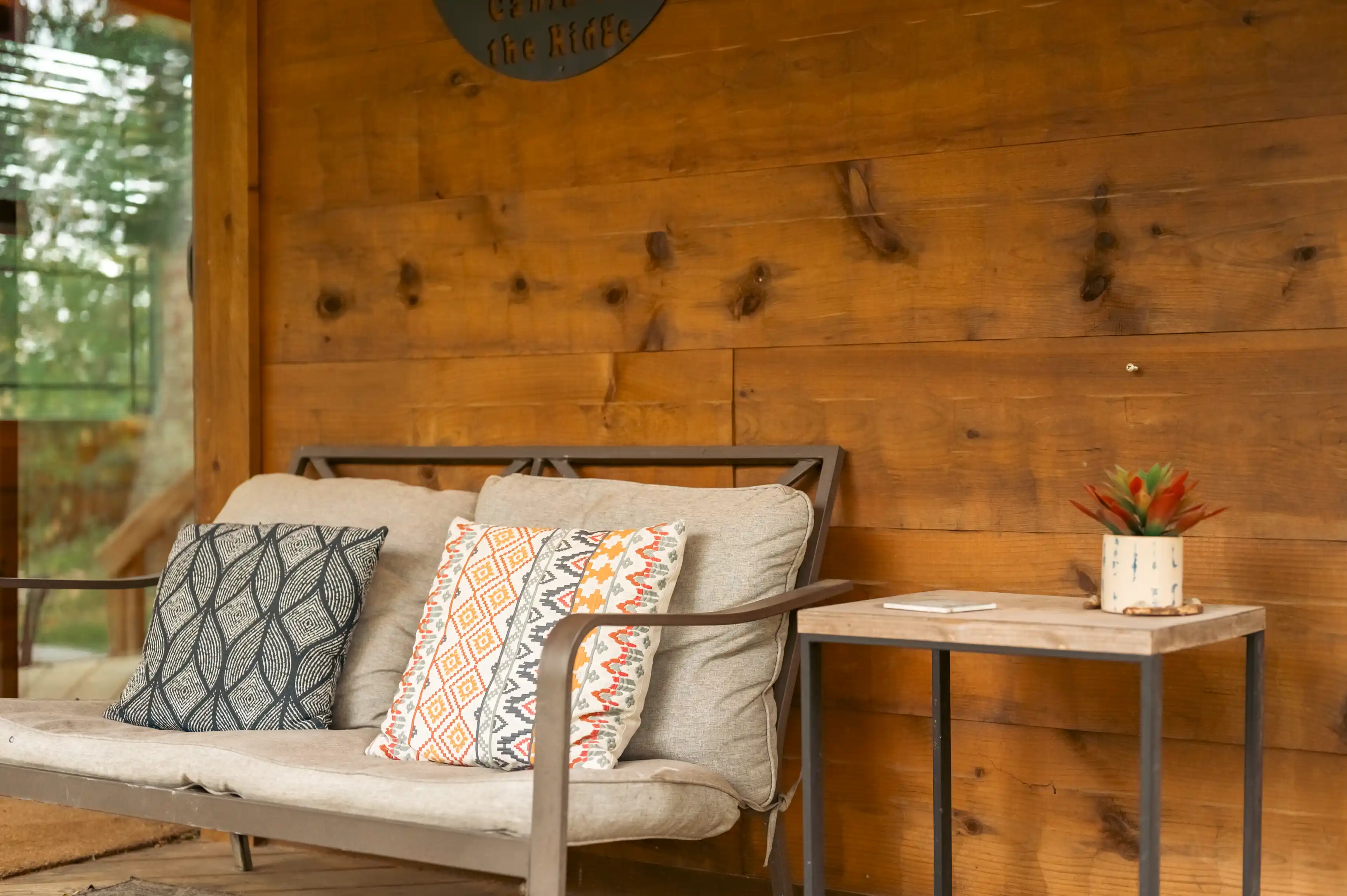 Cozy outdoor seating area with a modern sofa adorned with patterned pillows next to a wooden side table featuring a potted succulent plant, against a warm wooden cabin wall.