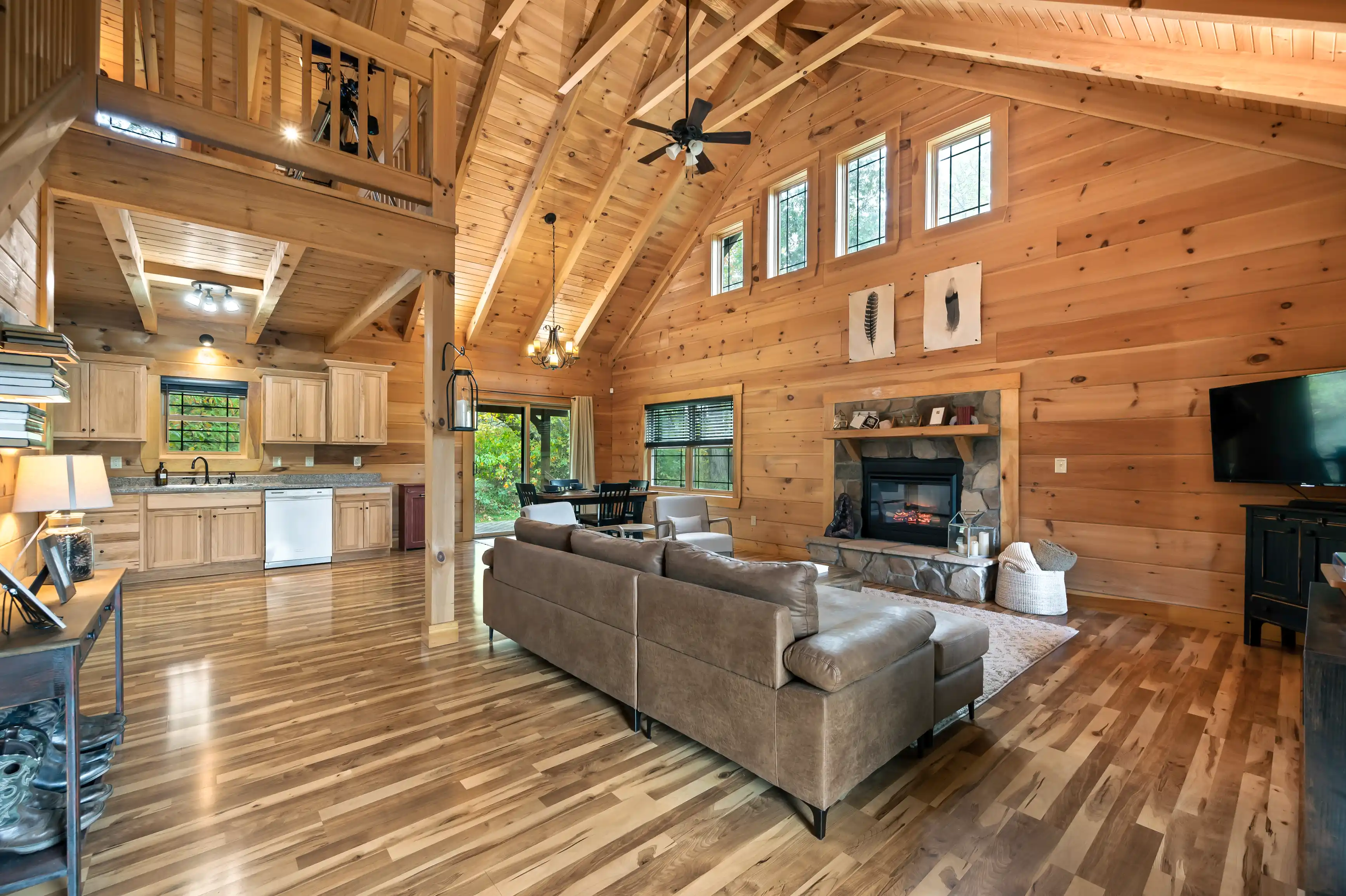 Spacious cabin interior with high vaulted ceilings, wood walls and floors, a fireplace, and a loft area, furnished with a sectional sofa, a dining set, and a kitchen with modern appliances.
