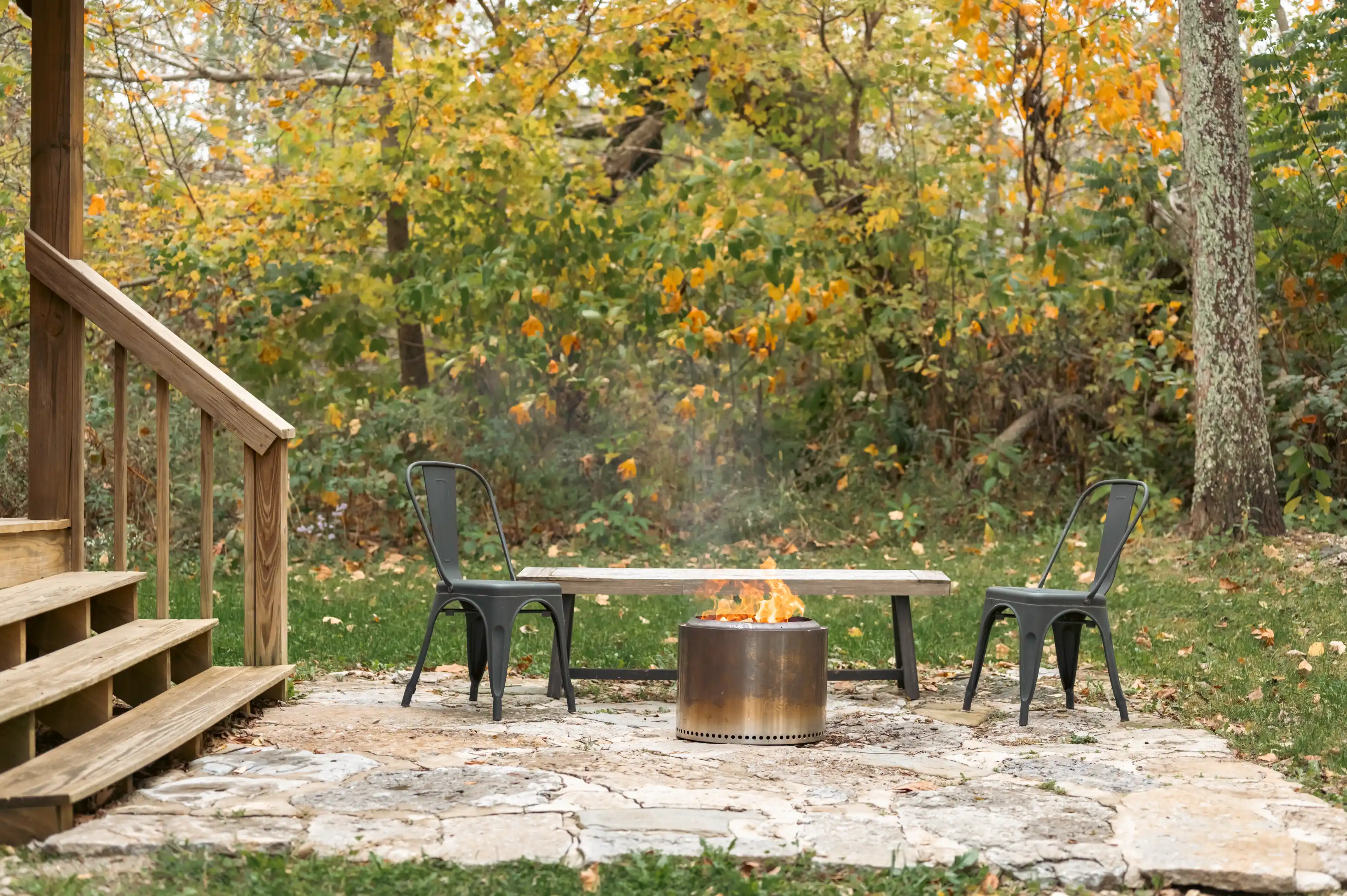 Cozy backyard setting with fire pit ablaze and two chairs by a wooden table on a flagstone patio, surrounded by autumn trees.