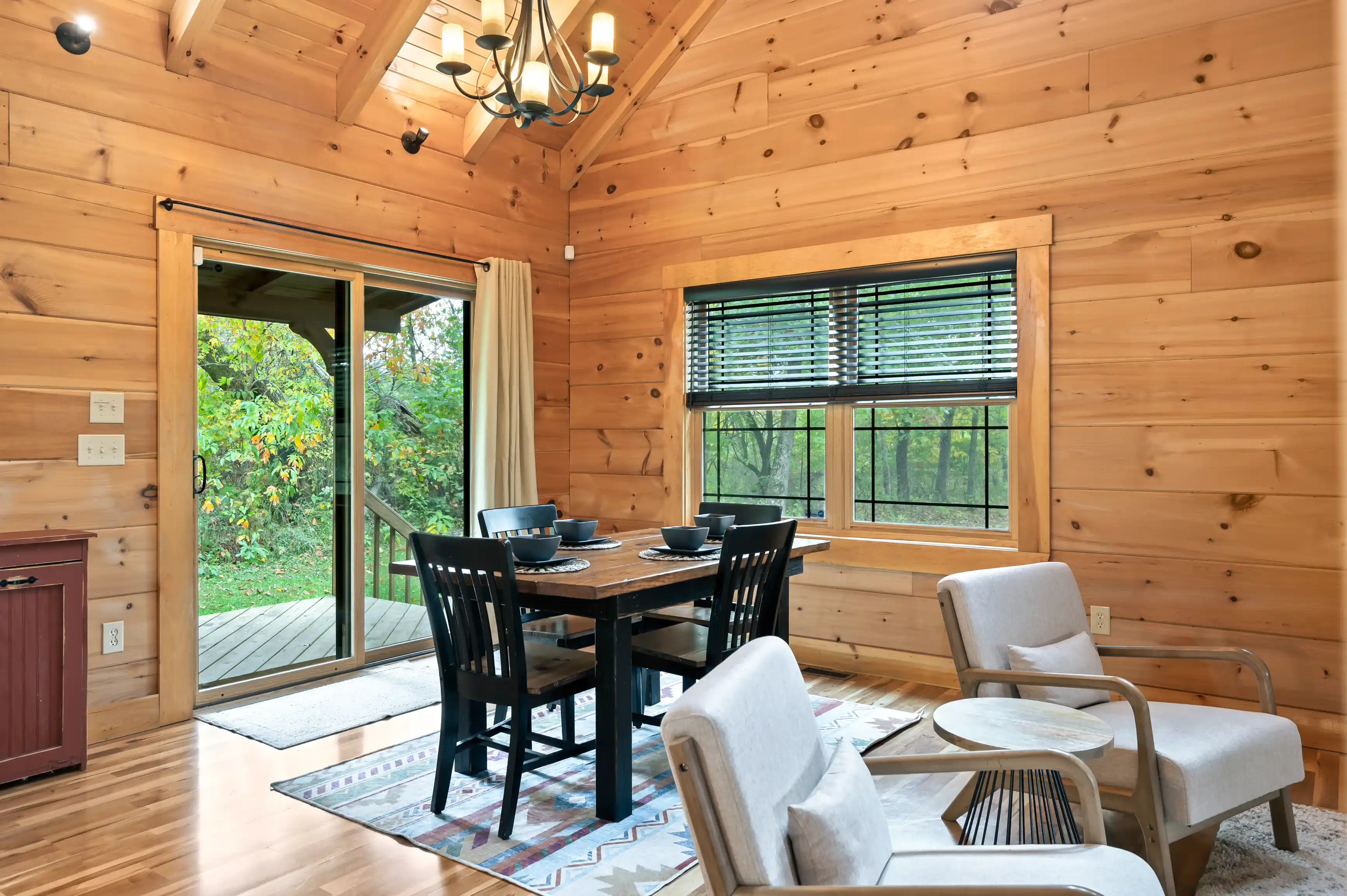 Cozy wooden cabin interior with dining table set, comfortable chairs, and a view of the woods through windows.
