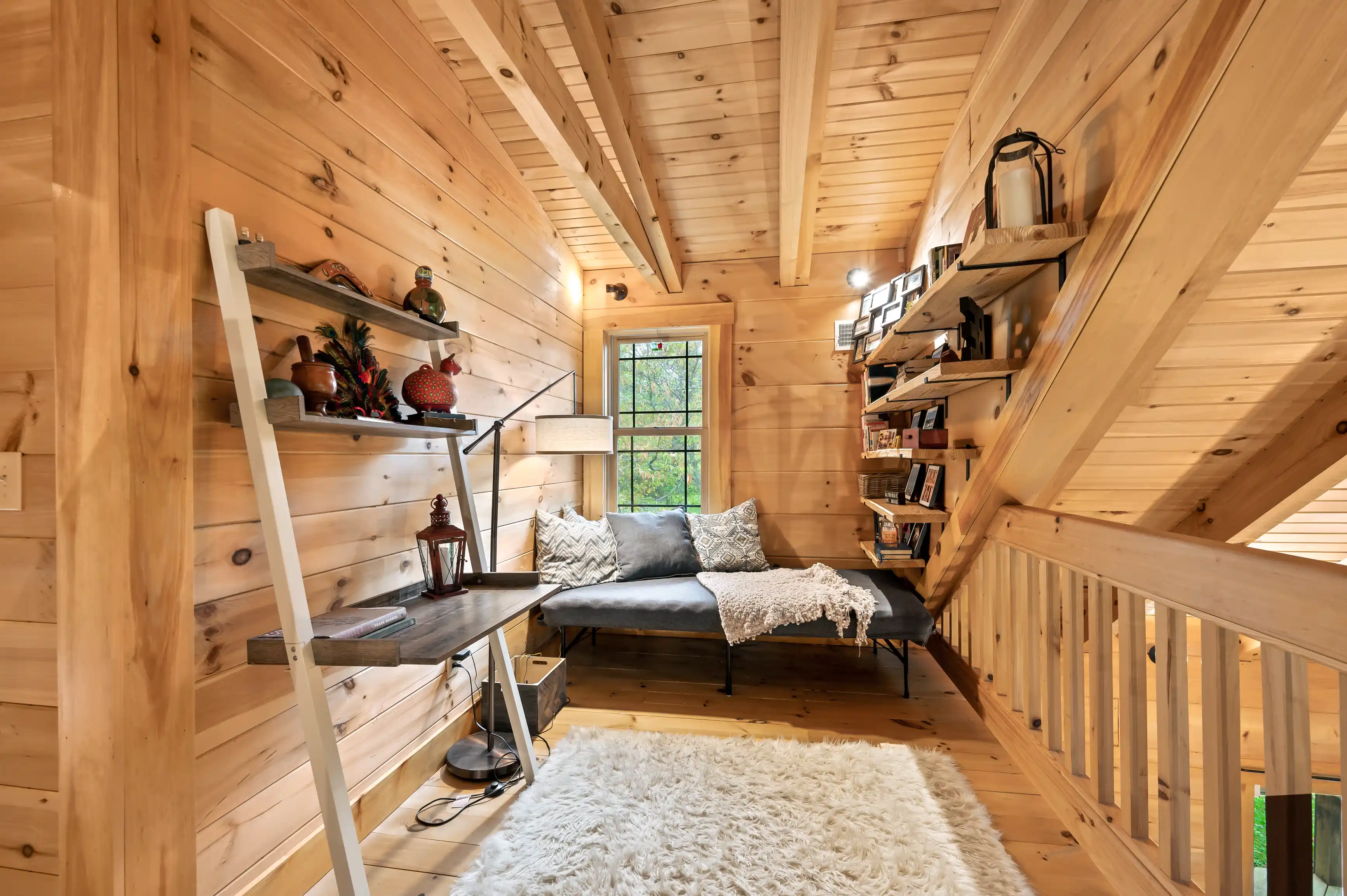 Cozy wooden interior of a cabin with a daybed, shelves, bookcase under stairs, and large window with nature view.