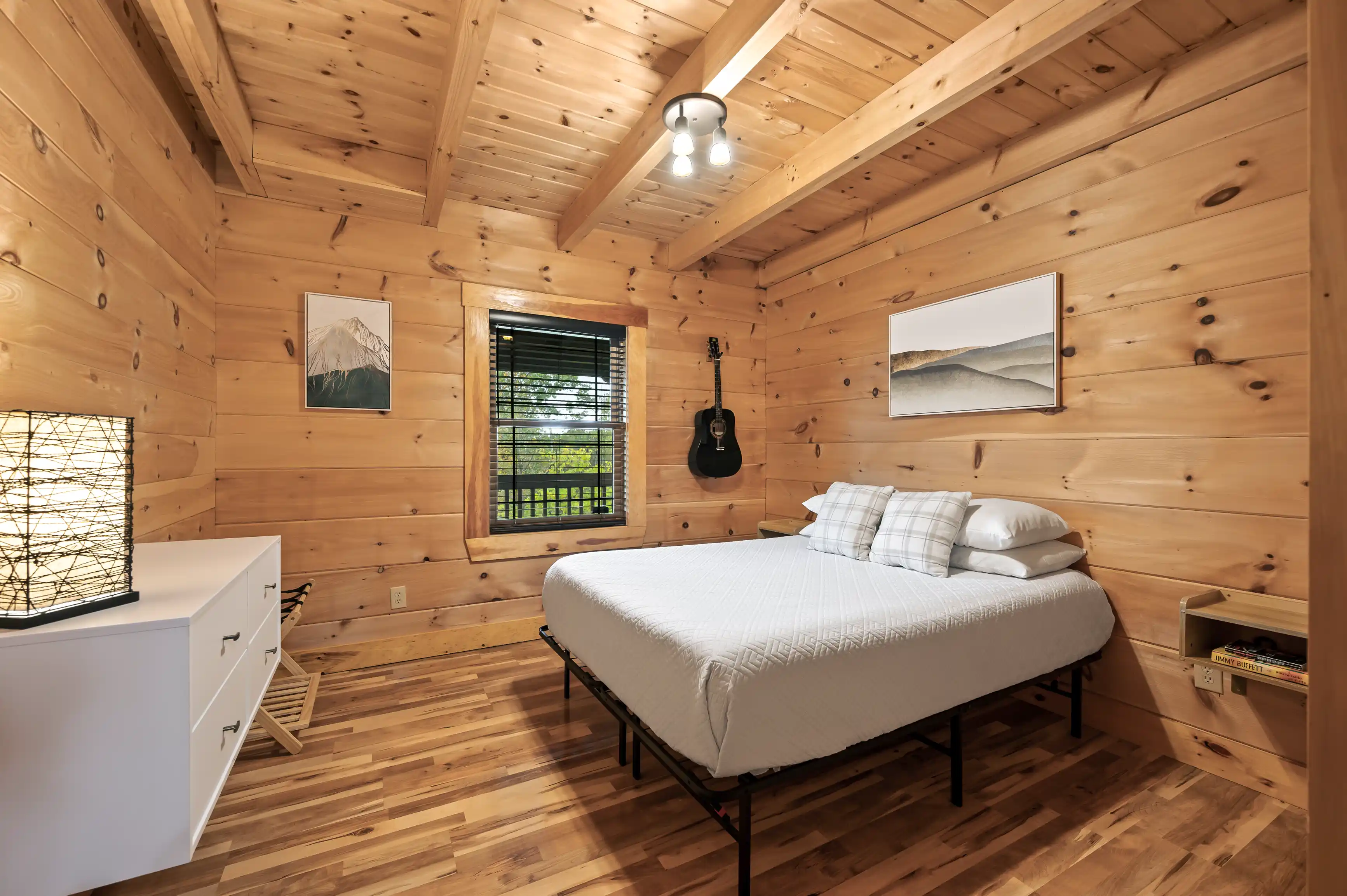 Cozy cabin bedroom interior with wooden walls and ceiling, queen-sized bed, framed artwork, wall-mounted guitar, and window with nature view.