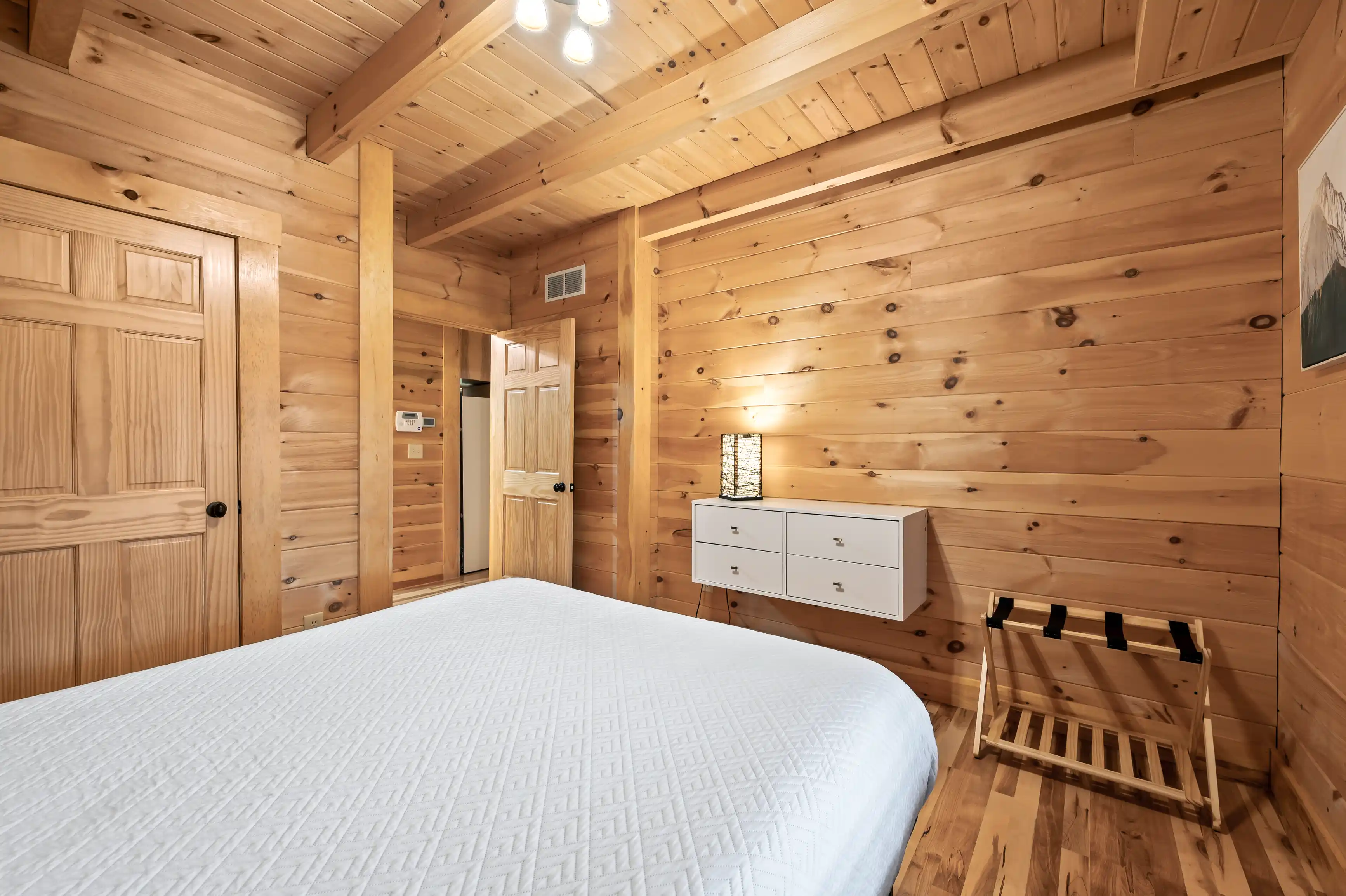 Cozy wooden cabin bedroom interior with a large bed, white bedding, natural wood walls, ceiling, and floors, complemented by modern light fixtures and simple furnishings.