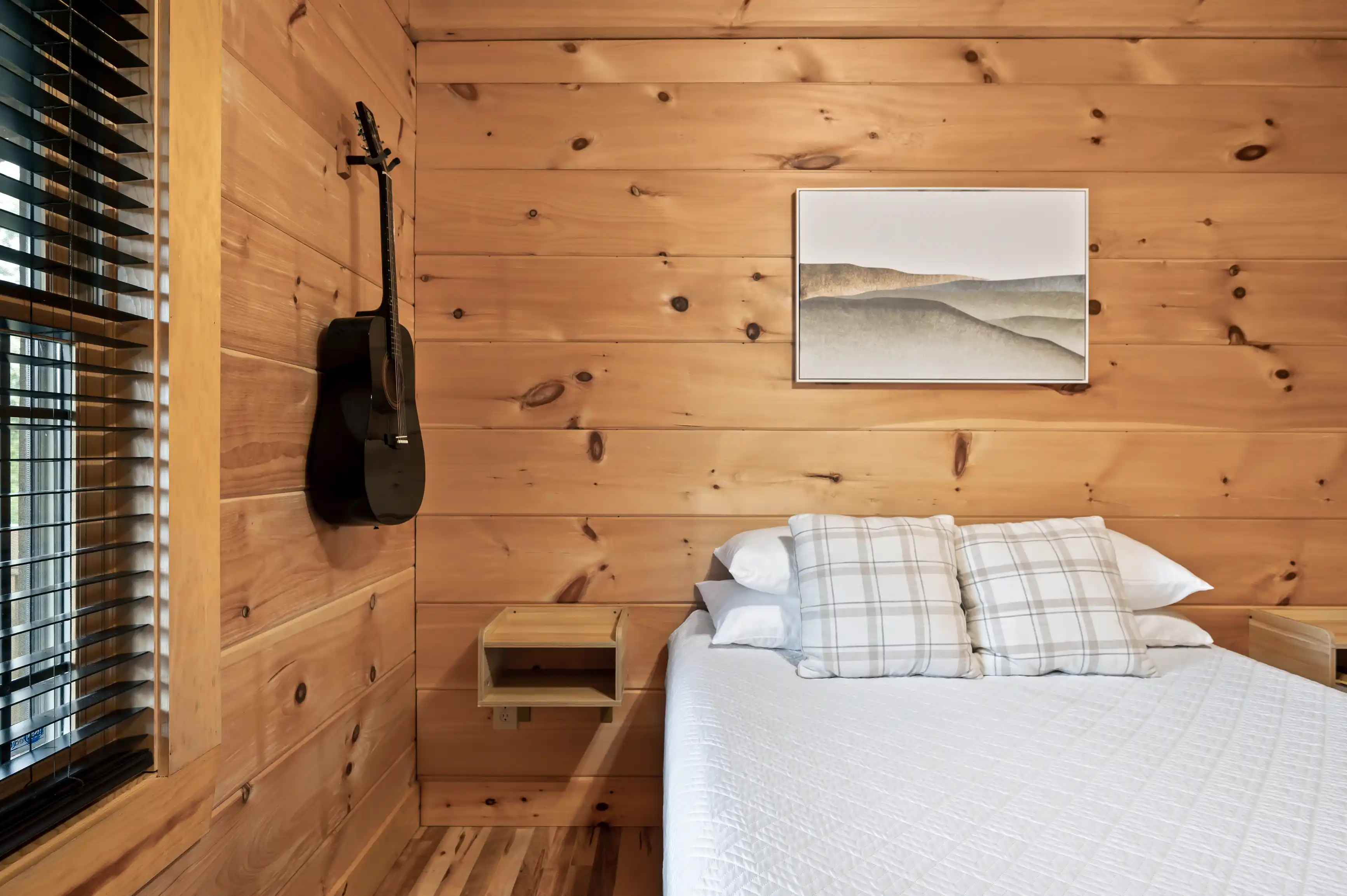 Cozy wooden cabin bedroom interior with a neatly made bed, plaid pillows, a black acoustic guitar on the wall, a framed landscape picture, and closed window blinds.