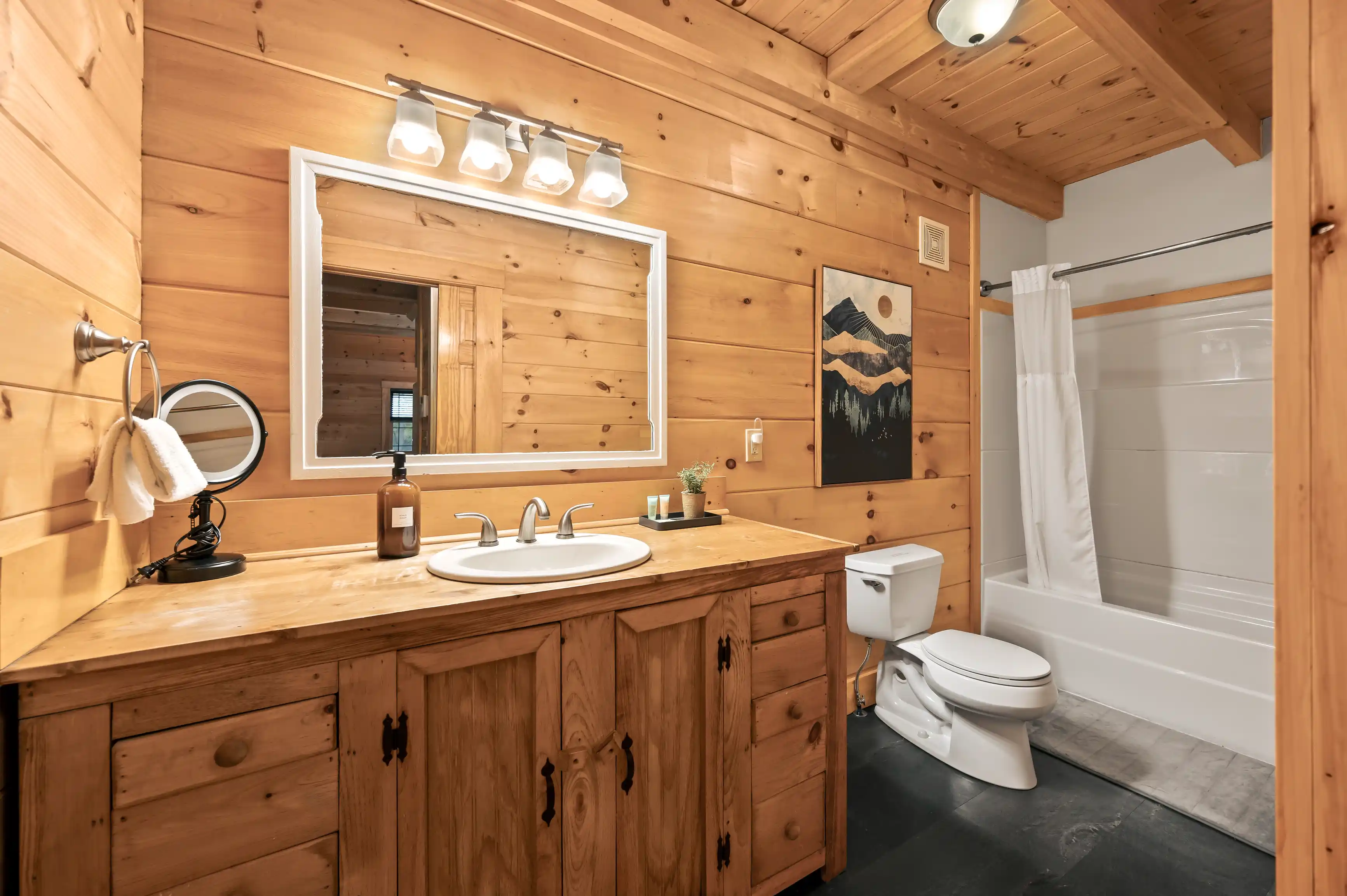 Rustic bathroom interior with wooden walls and cabinets, featuring a large mirror, double sink vanity, wall-mounted lights, magnifying mirror, toilet, and shower with a white curtain. A mountain landscape picture hangs on the wall.