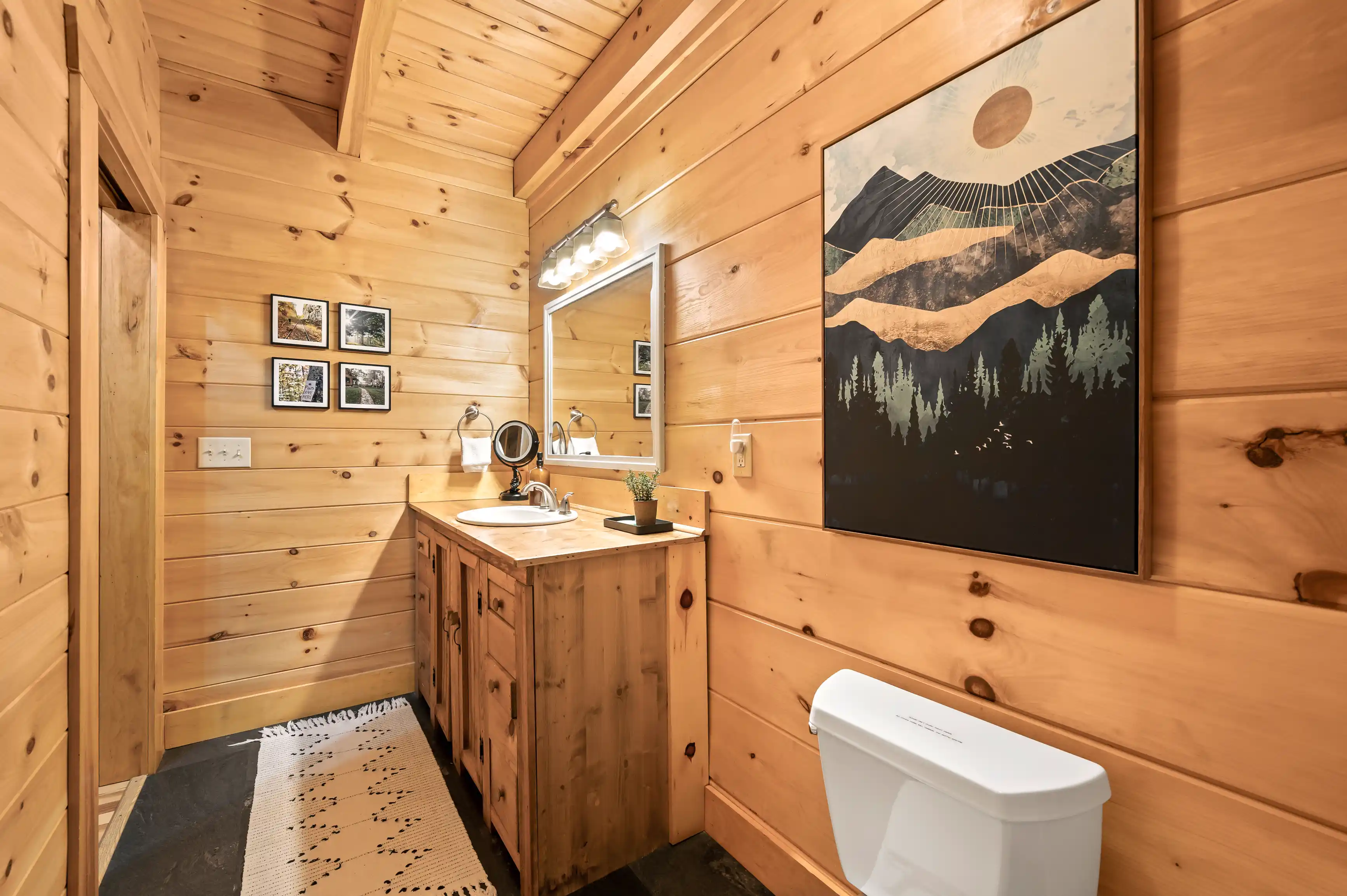Rustic bathroom interior with wooden walls and floors, a wooden vanity with a basin, a mirror, a toilet, and decorative art.