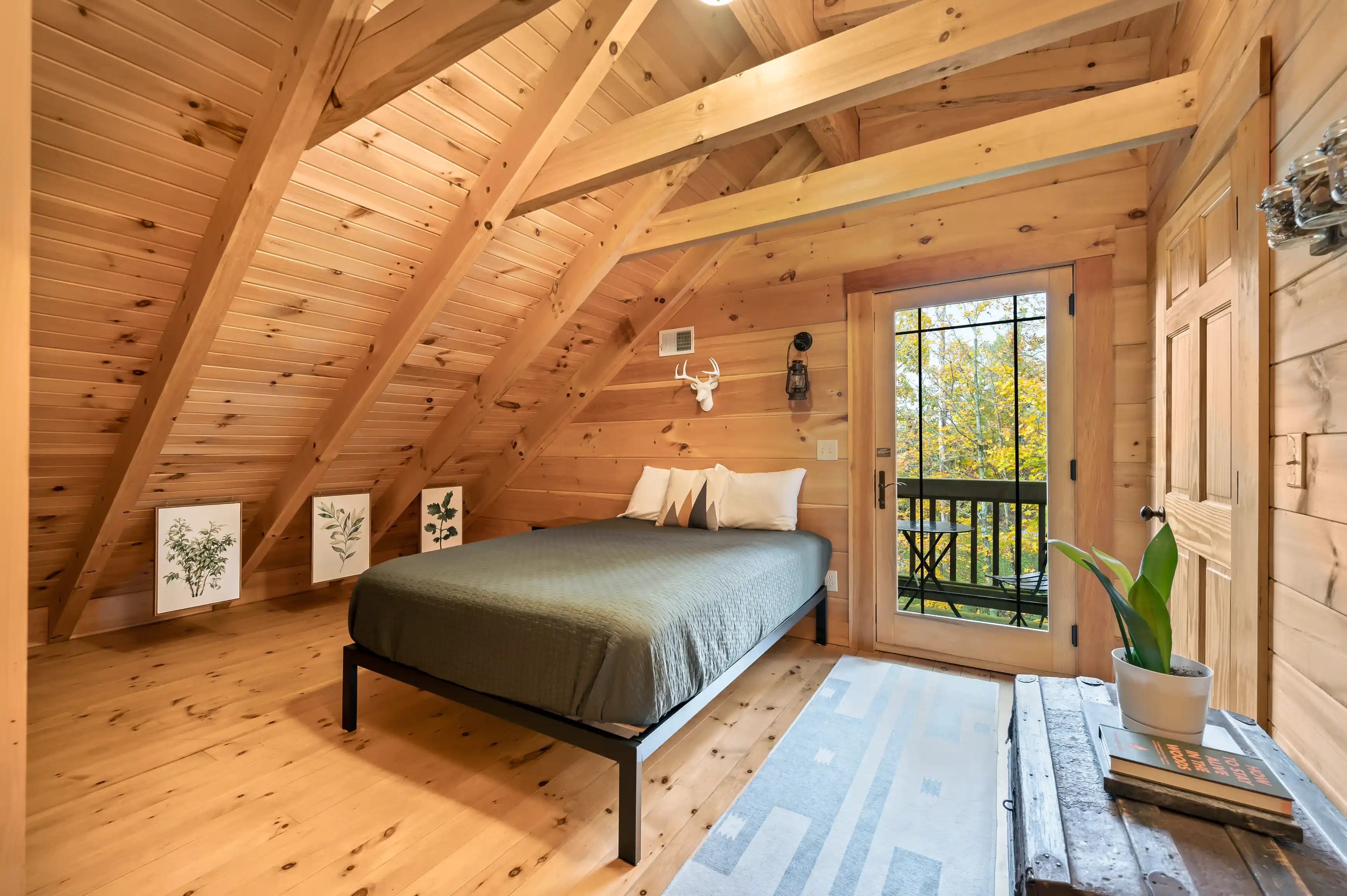 A cozy wooden cabin bedroom with a slanted ceiling, a double bed, white bedding, framed botanical art on the walls, and a balcony with a forest view visible through the glass door.