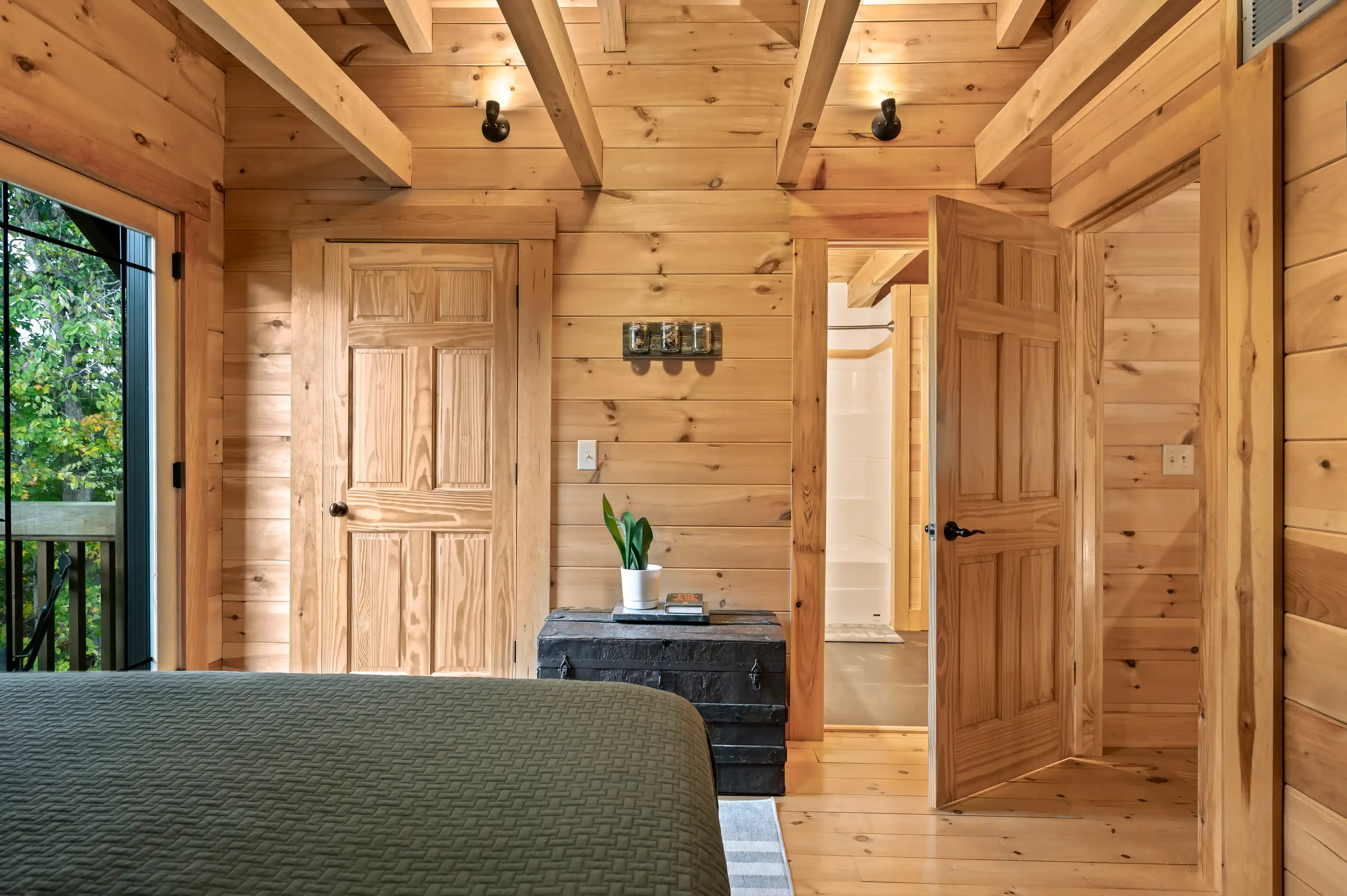 Interior view of a cozy cabin bedroom with wooden walls, a glimpse of a bed, open doors to a bathroom, and natural light from a glass door leading to a balcony.