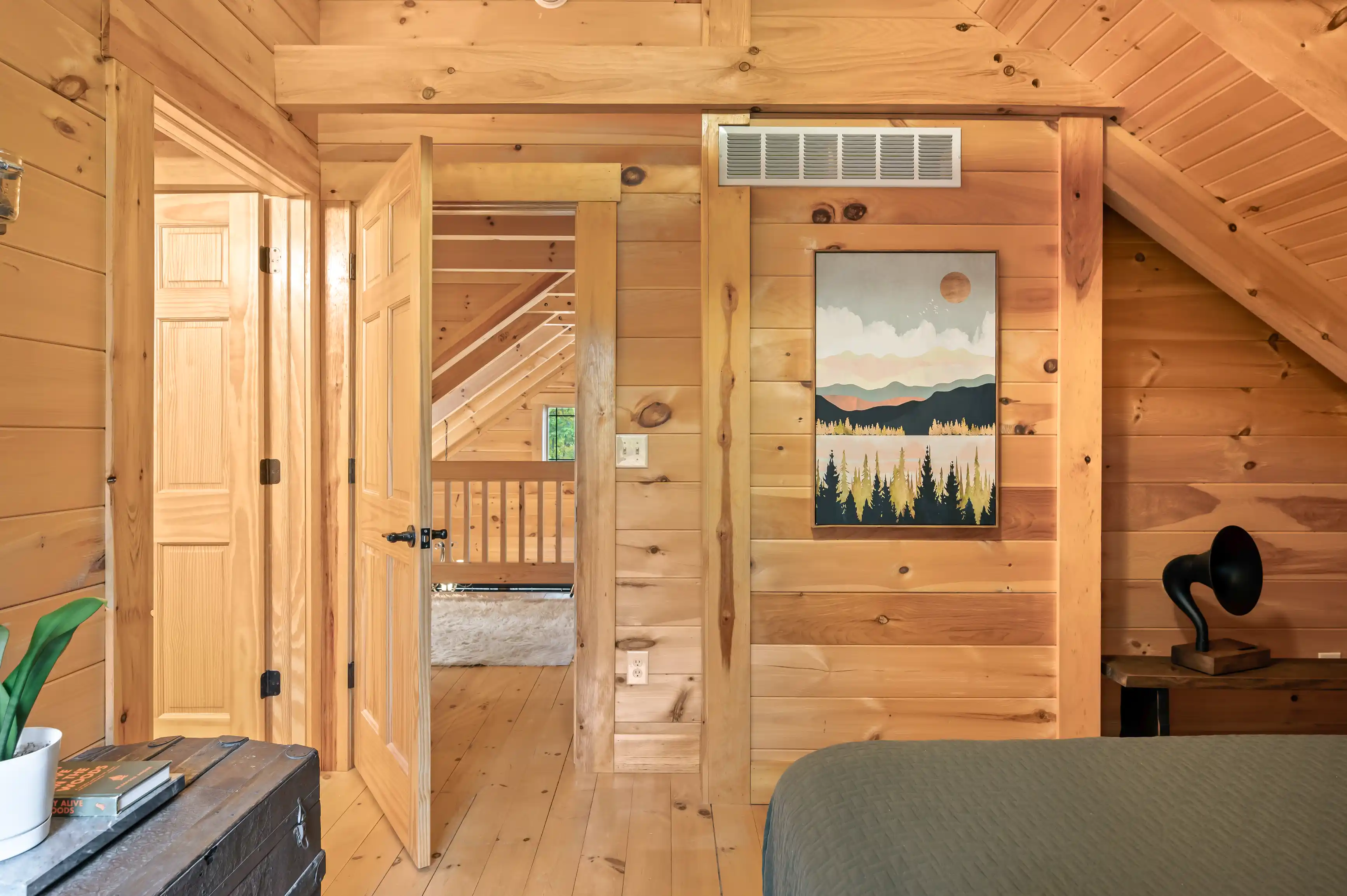 Interior of a cozy cabin with wooden walls, floors, and ceiling, featuring an open door leading to a balcony, a framed landscape painting, and a bed with a grey cover.