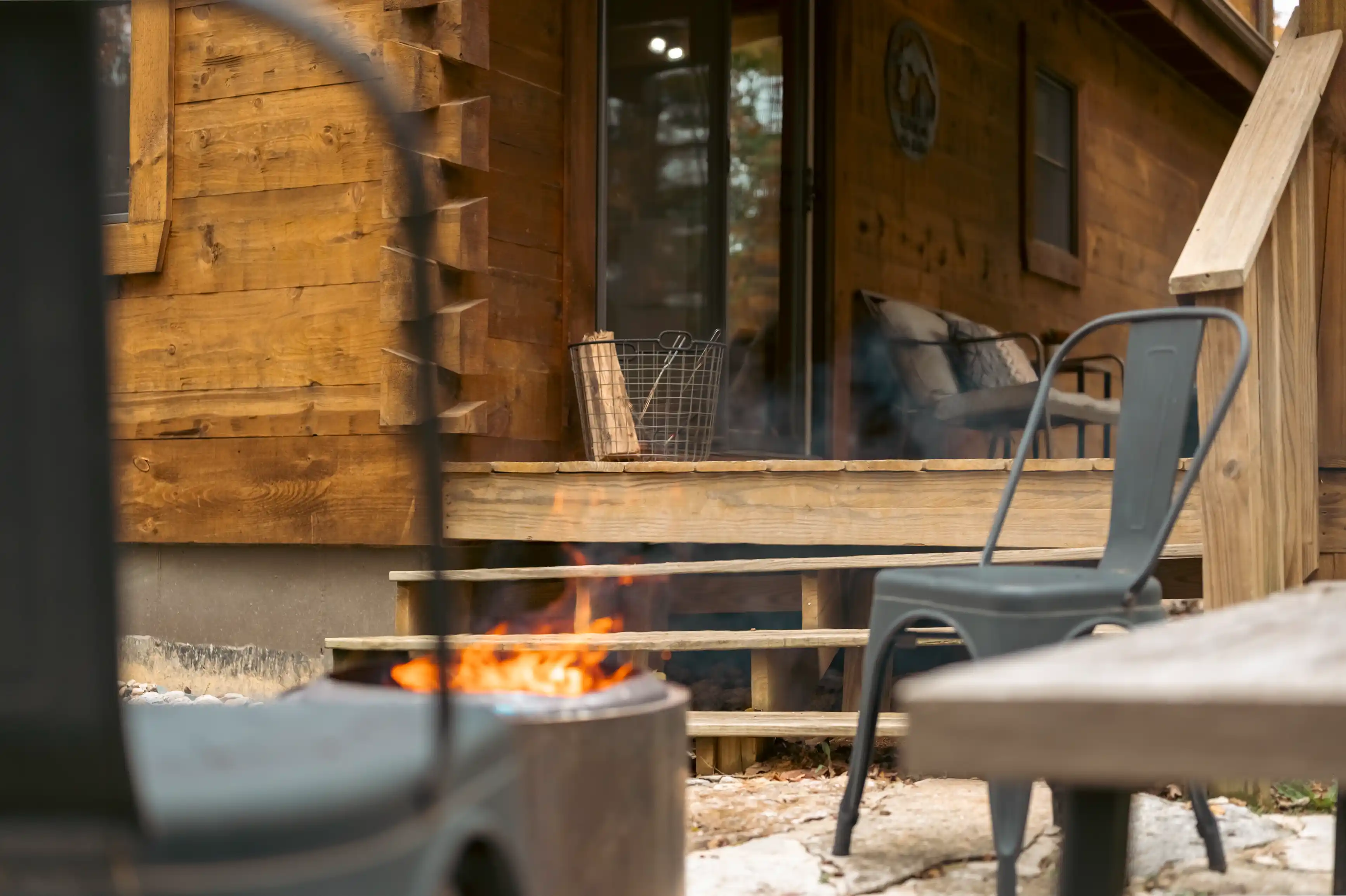 Cozy wooden cabin porch with a lit fire pit in focus and two metal chairs around it.