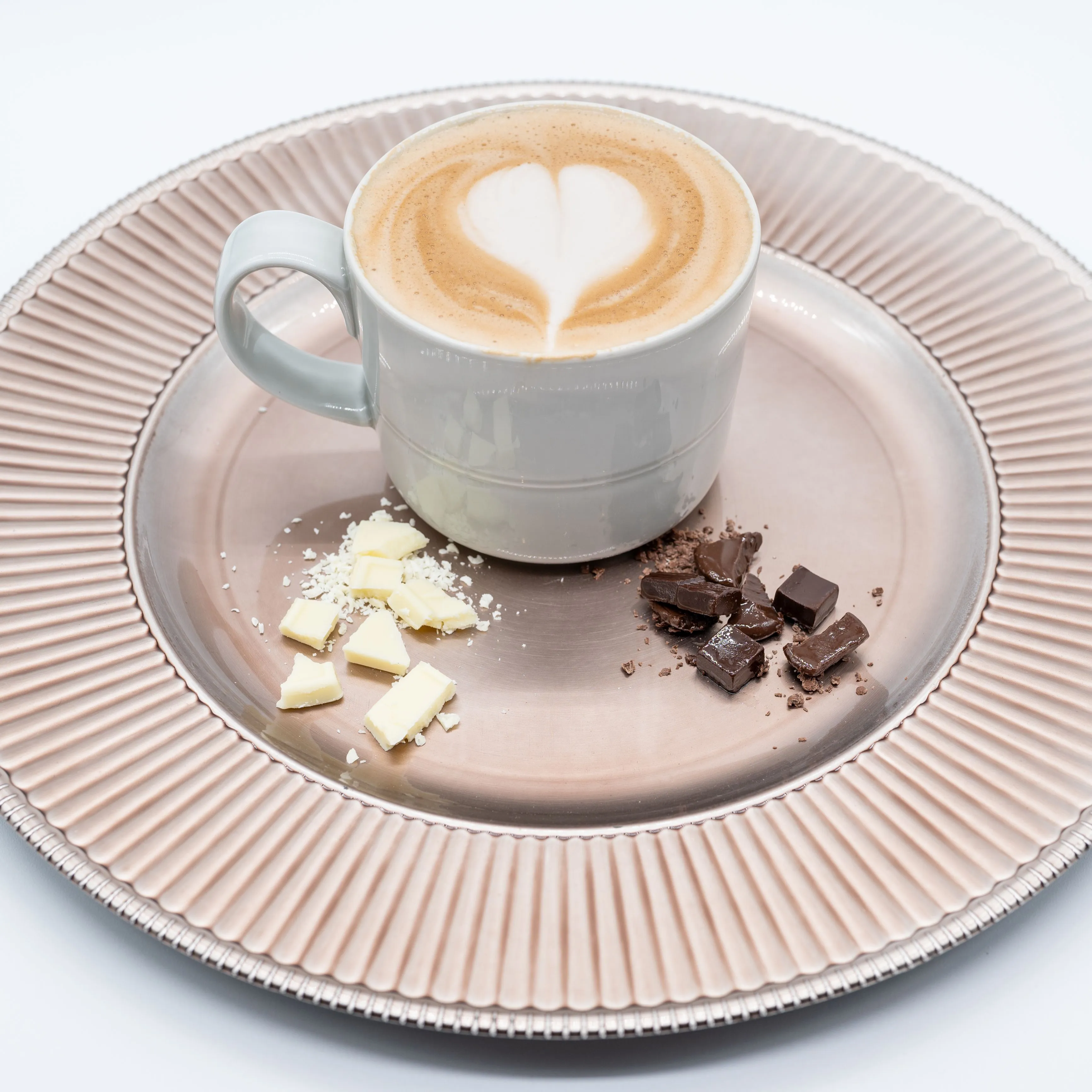 A cup of cappuccino with heart-shaped latte art on a ribbed bronze plate with pieces of white and dark chocolate.