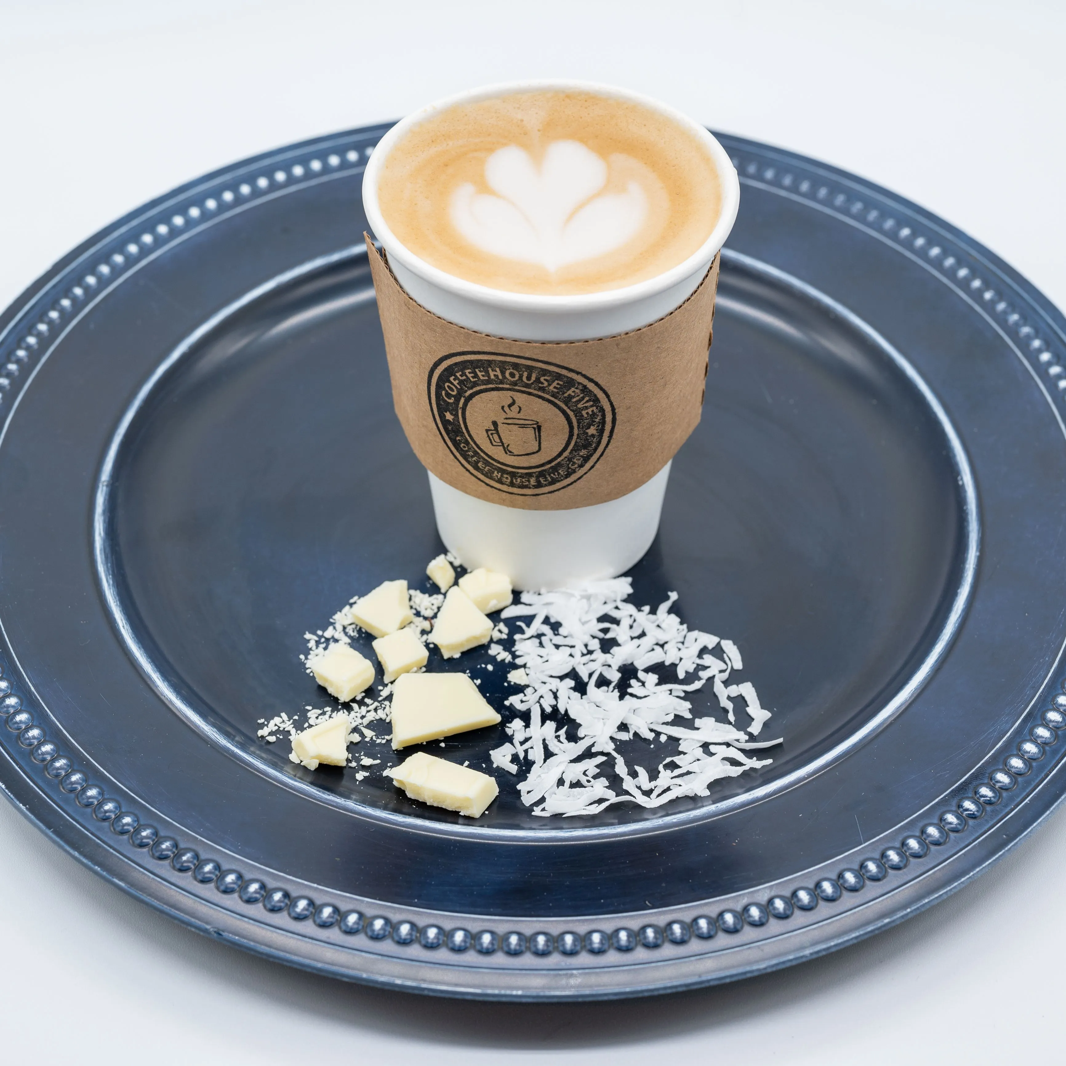 A paper cup of cappuccino with latte art on top, placed on a navy blue plate accompanied by a few pieces of white chocolate and shredded coconut, isolated on a white background.