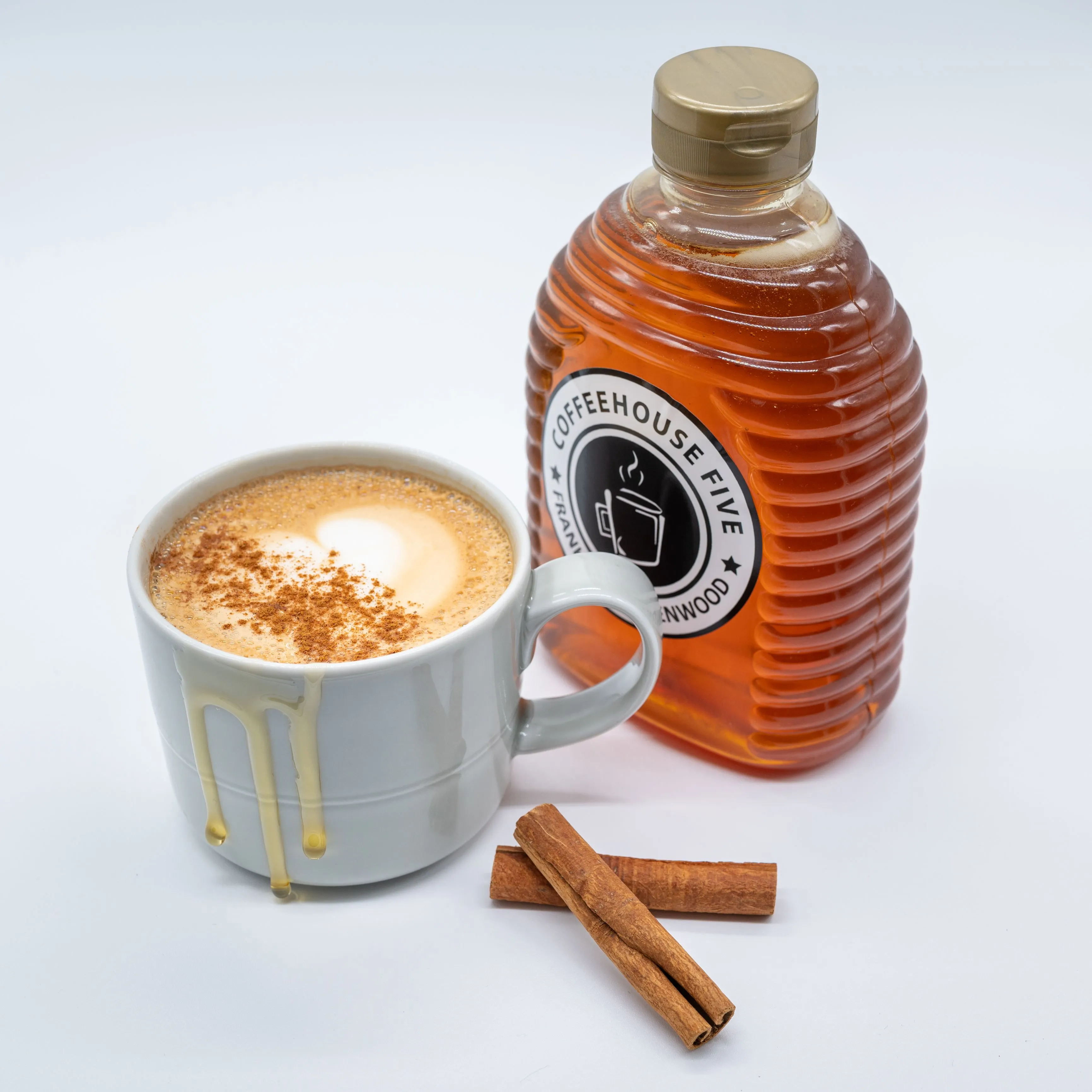 A cup of cappuccino with heart-shaped foam art next to a large bottle of Coffeehouse Five syrup and two cinnamon sticks on a white background.