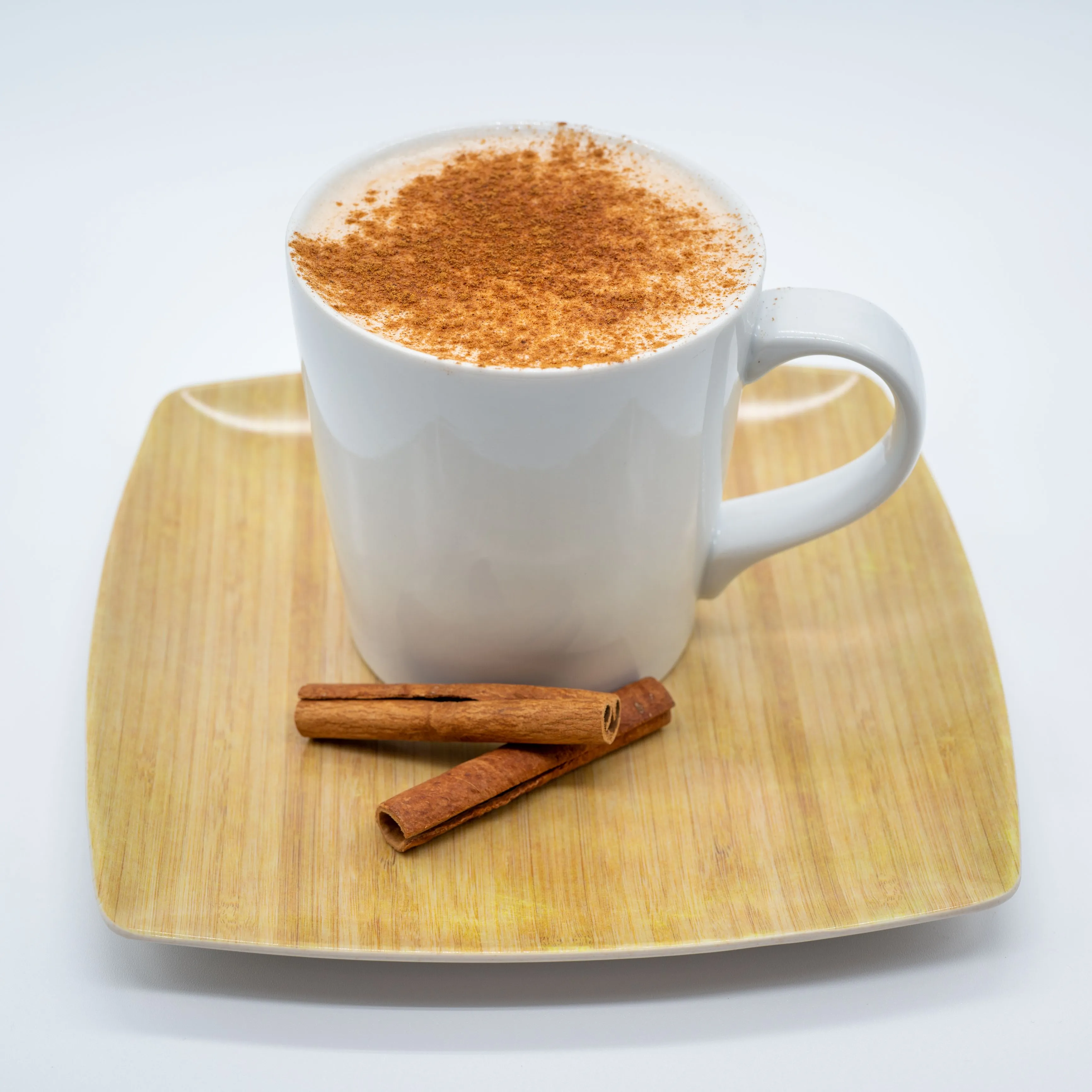A cup of cappuccino with cinnamon sprinkled on top, resting on a wooden tray beside two cinnamon sticks.
