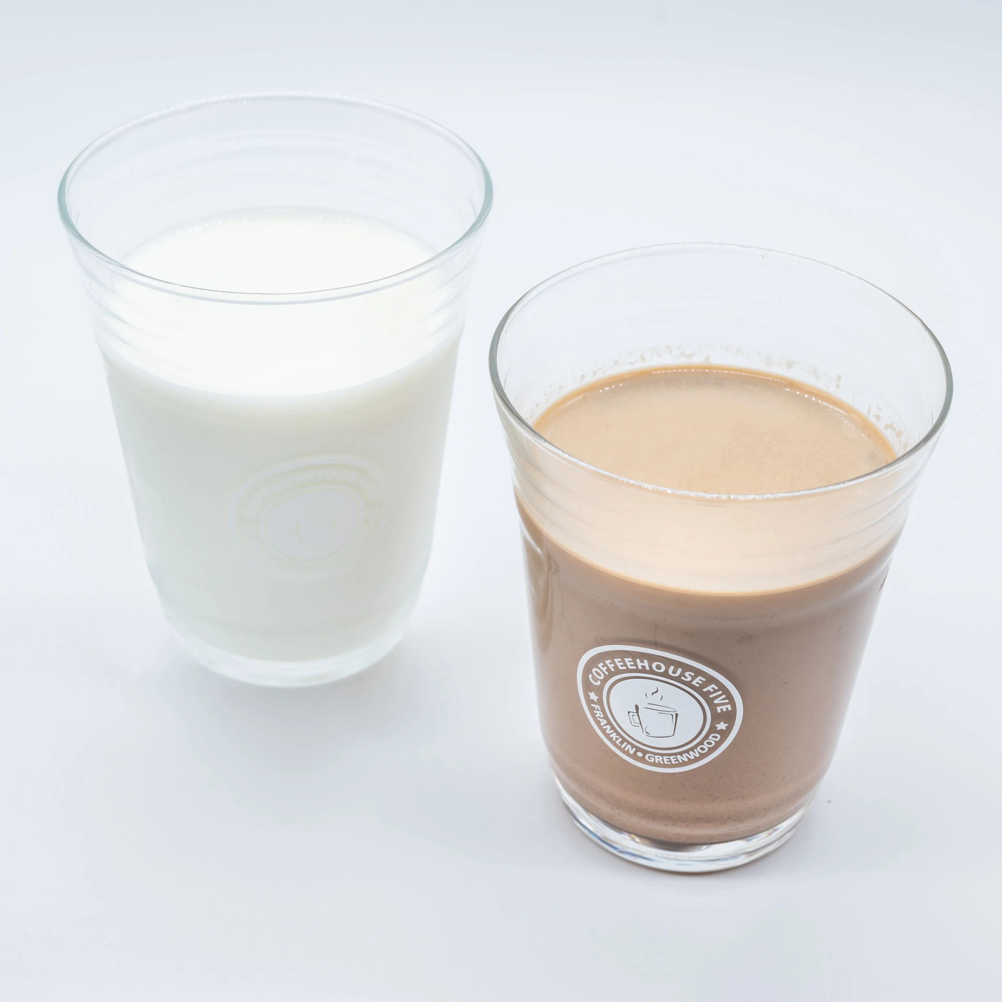 Two glasses on a white surface, one with milk and the other with a chocolate-flavored beverage, both with printed logos.
