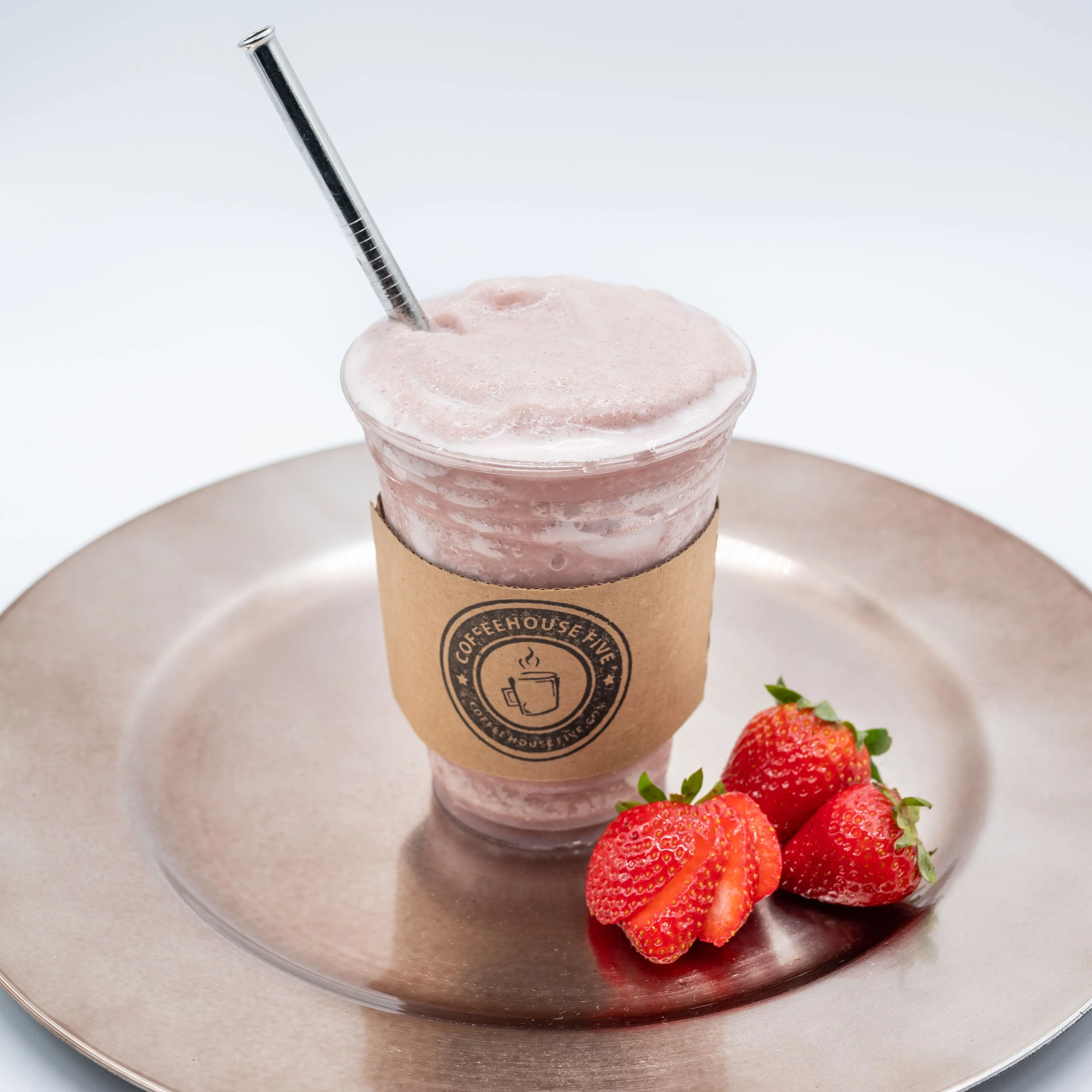 A strawberry smoothie in a clear plastic cup with a metal straw, branded paper sleeve around the cup, on a bronze plate with fresh strawberries beside it, against a white background.