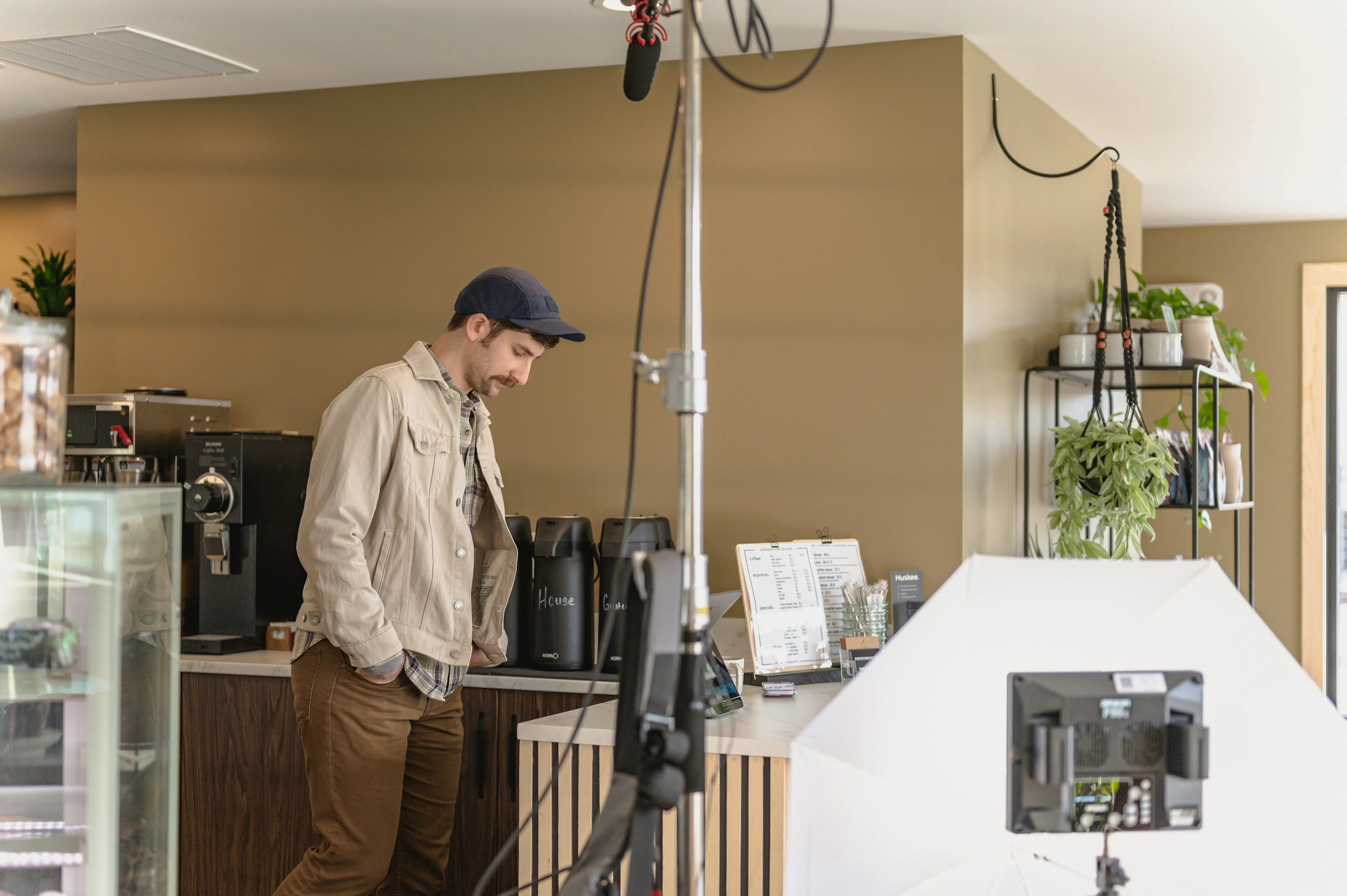 Man in casual clothing standing pensively in a cafe beside a counter with coffee equipment, surrounded by filming gear including a camera and lighting equipment.