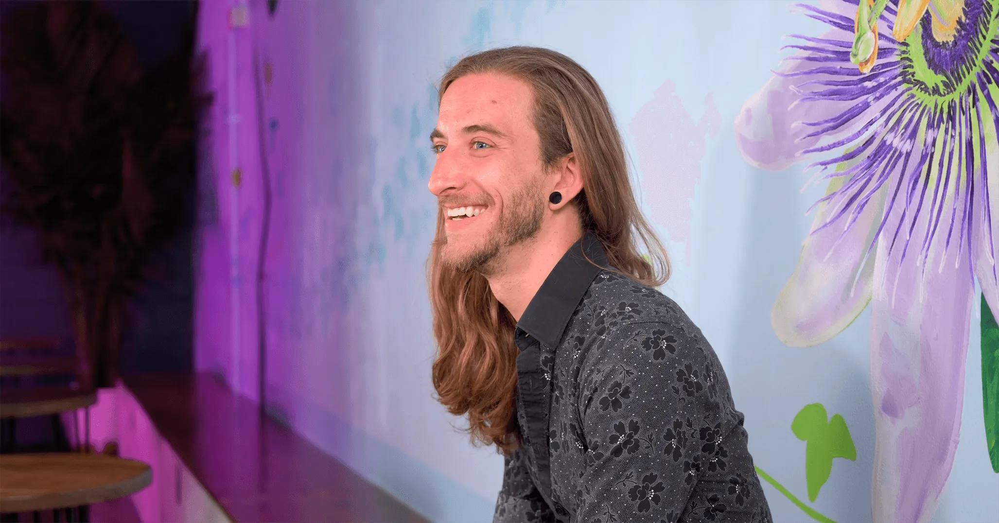 Side profile of a smiling man with long hair in front of a colorful mural with a large flower illustration.