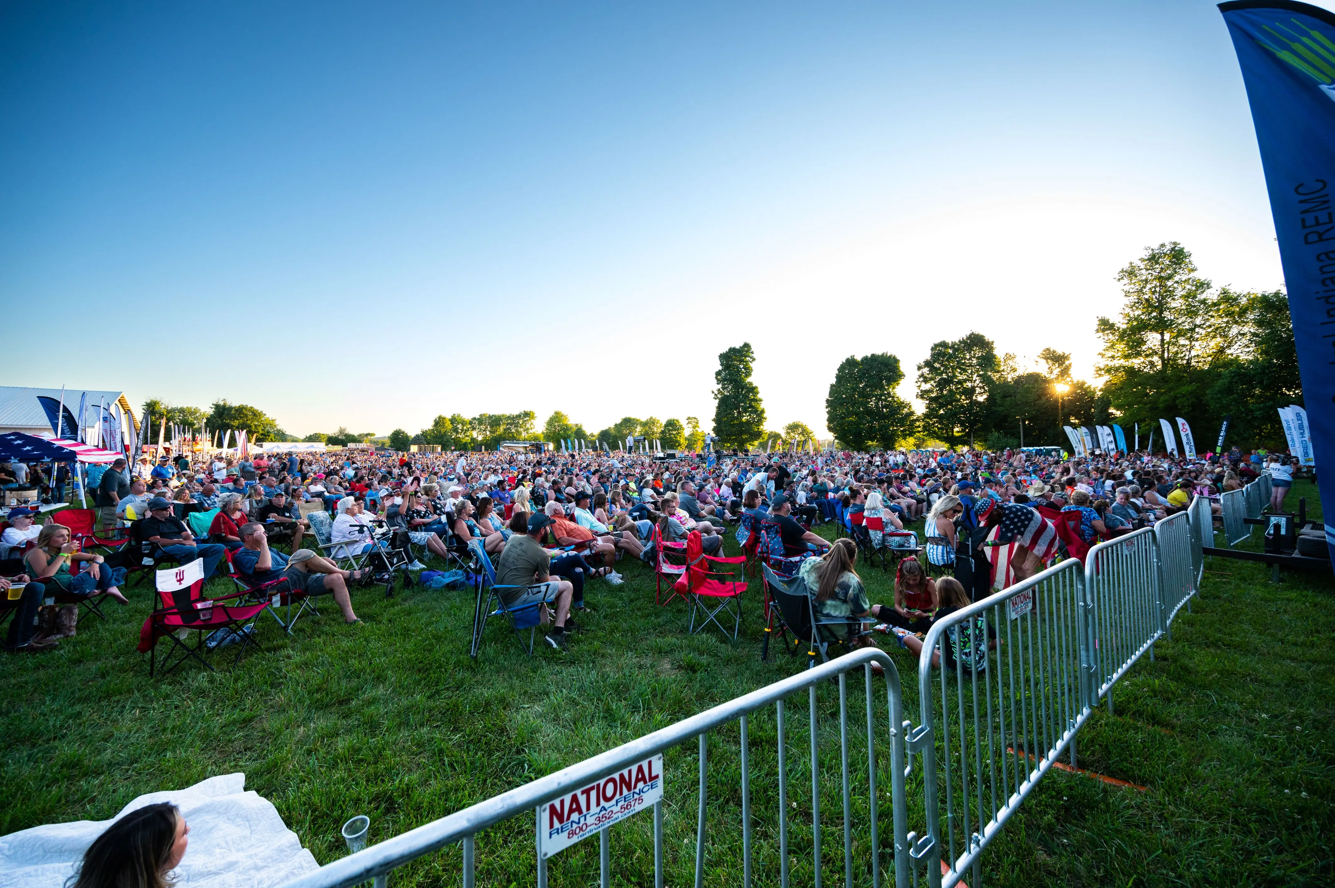 Outdoor event with a large crowd of people sitting on the grass behind barrier fences under a clear blue sky.