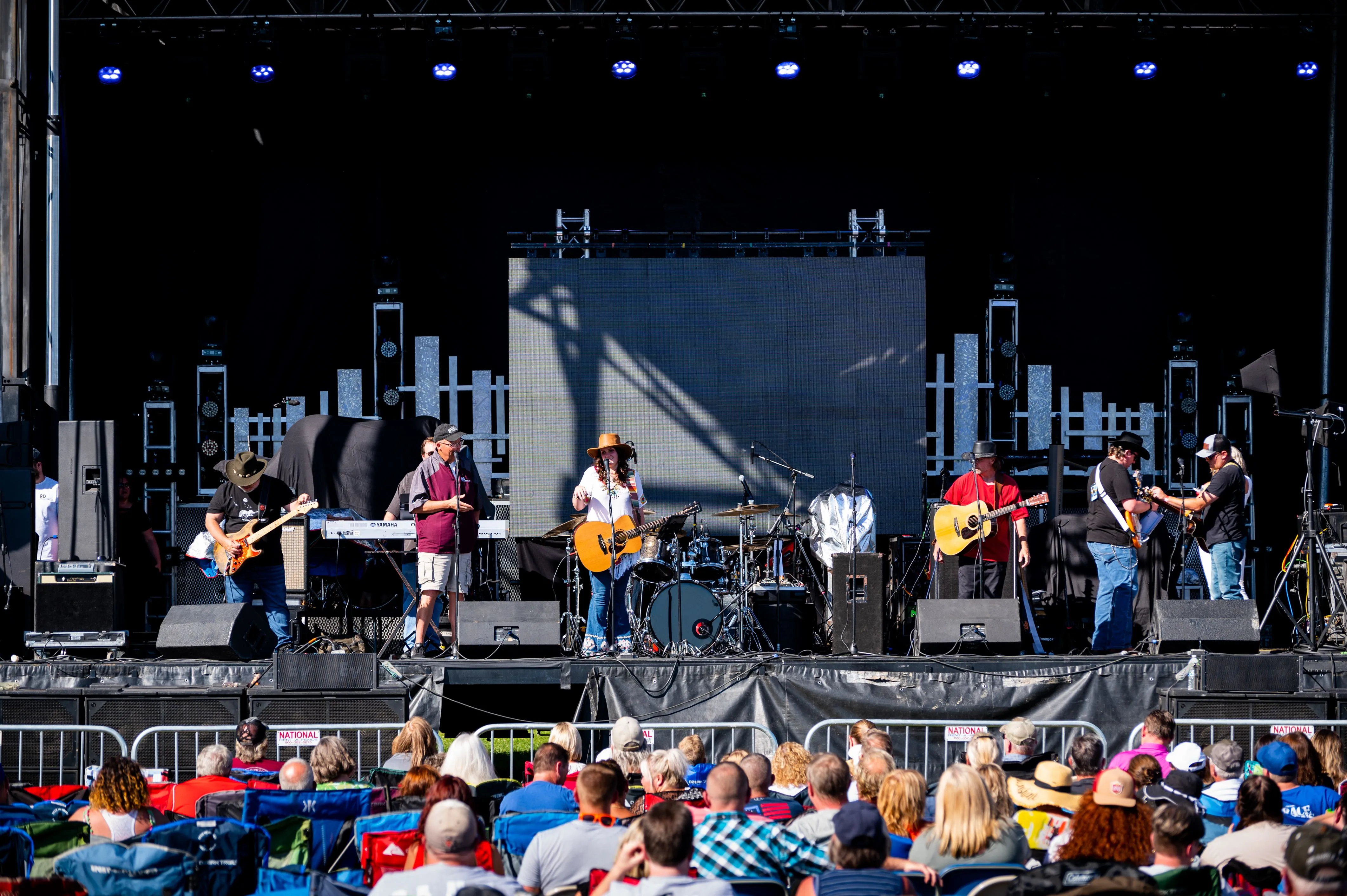 Band performing on an outdoor stage at a concert with an audience in the foreground sitting on folding chairs.