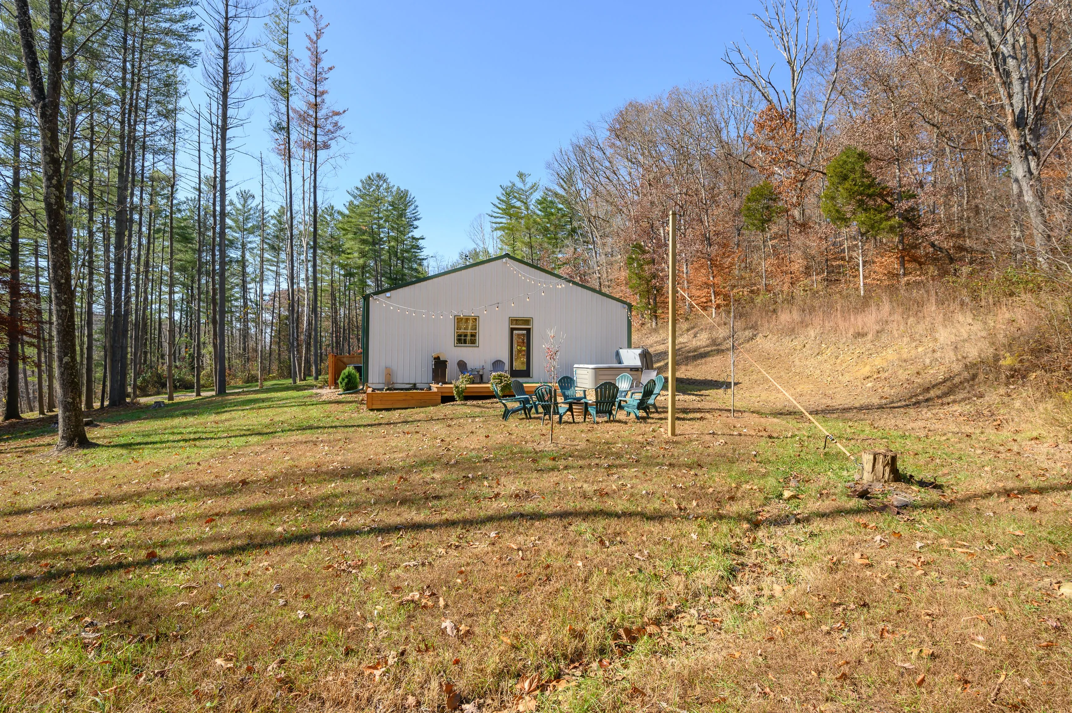 A small metal building with patio furniture outside located in a grassy clearing surrounded by forest with trees showing autumn foliage.