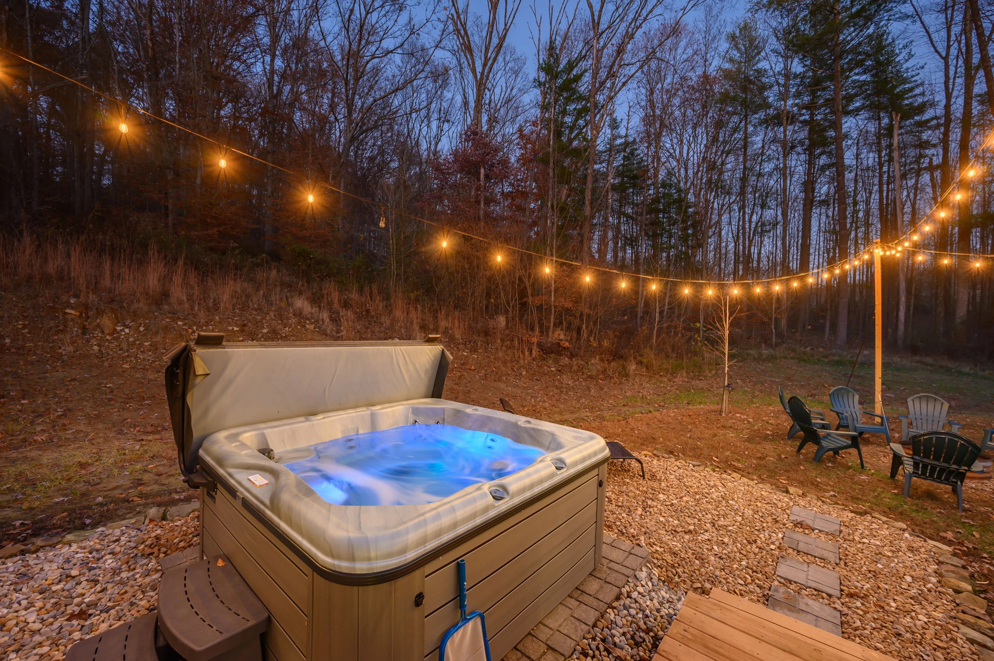 Outdoor hot tub with illuminated water, surrounded by string lights in a wooded backyard at dusk.
