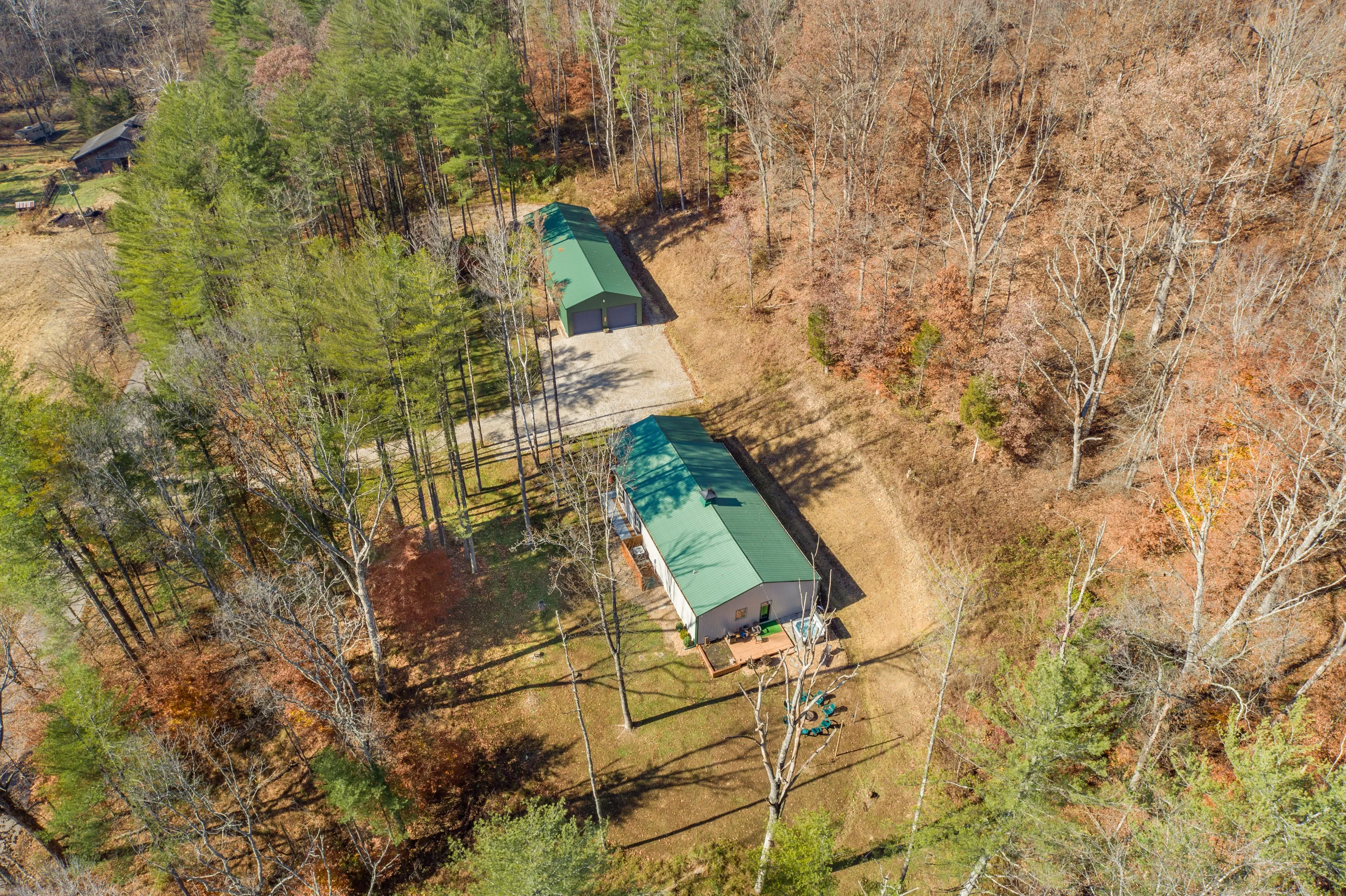 Aerial view of a rural property with two green-roofed buildings surrounded by a mix of coniferous and leafless deciduous trees with a playground in the foreground.
