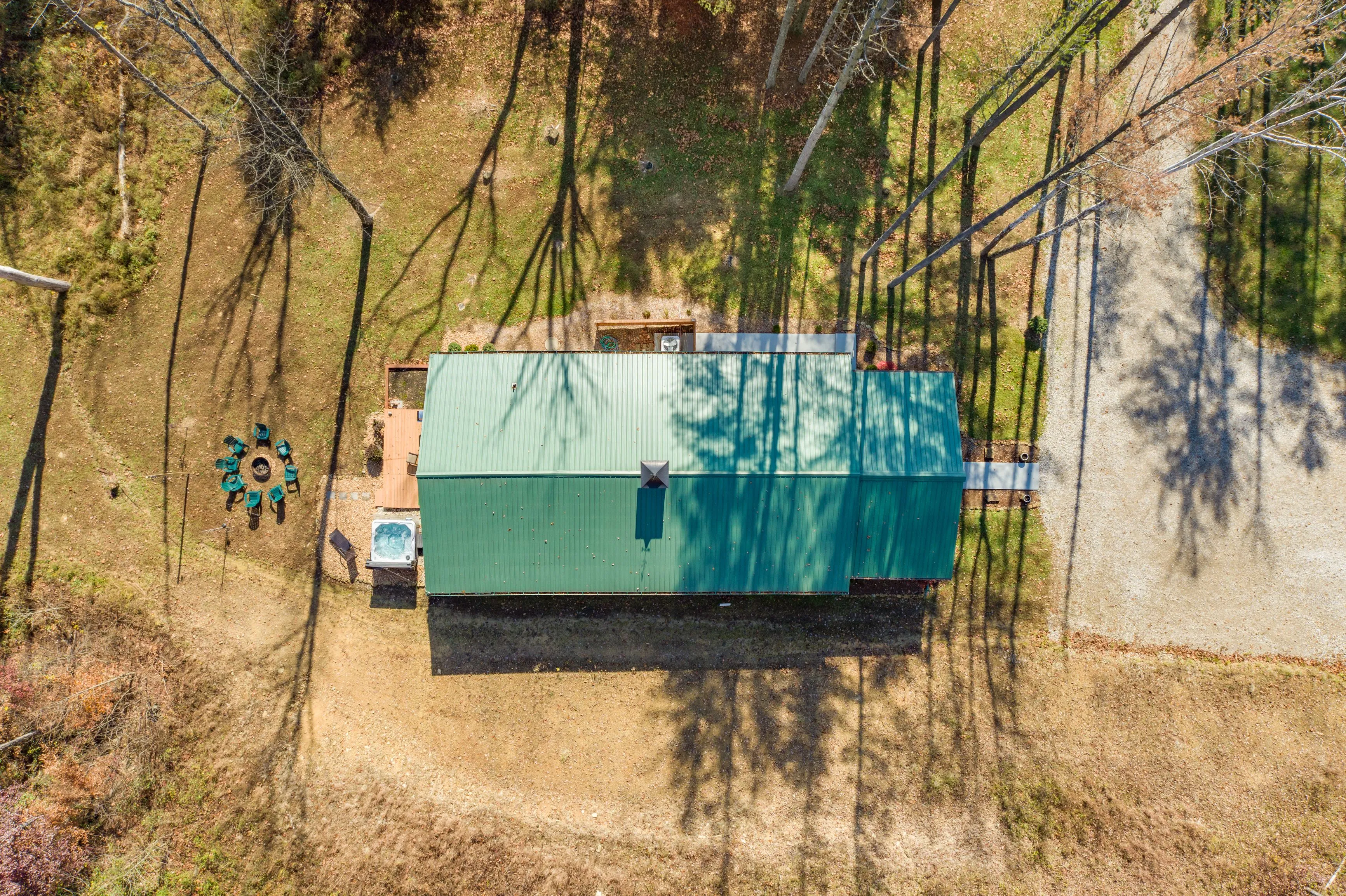 Aerial view of a green-roofed house with an adjacent hot tub and patio furniture surrounded by tall trees casting shadows on the ground.