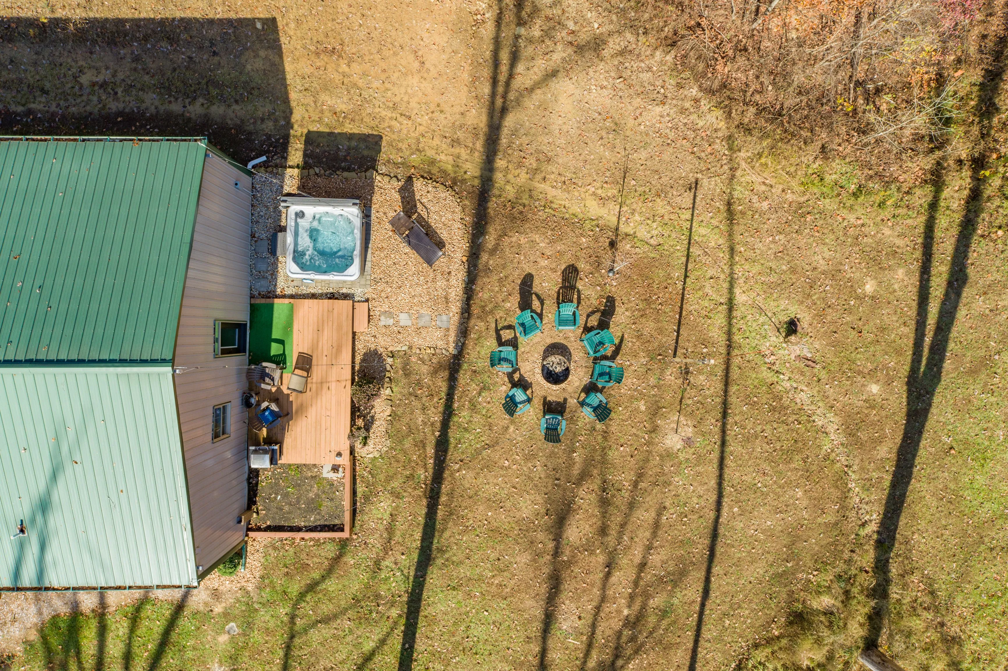Aerial view of a backyard with a green-roofed house, wooden deck, hot tub, and a circular arrangement of chairs around a fire pit.